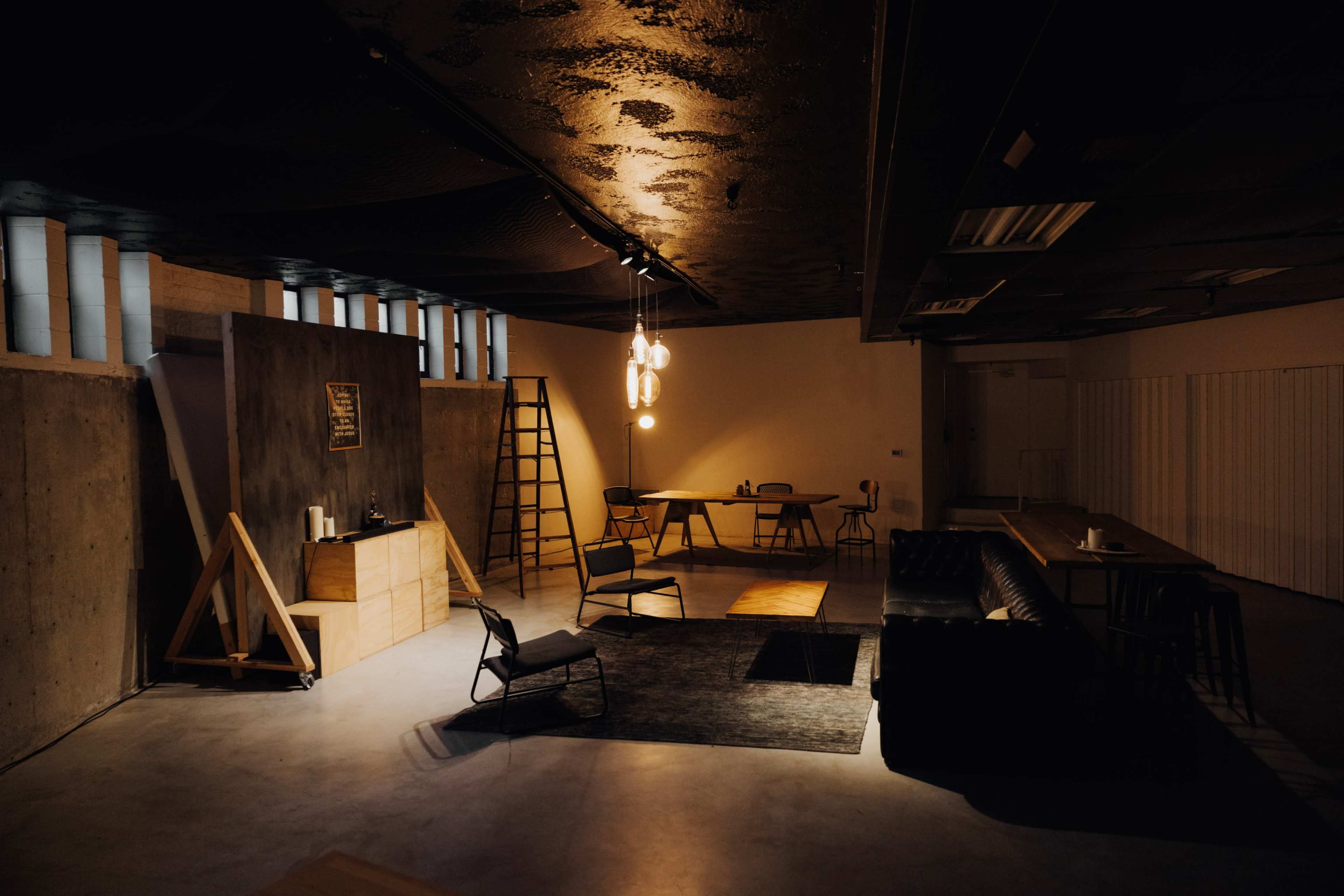 The image shows a dimly lit industrial-style room with concrete walls, modern furniture, and a mix of seating areas arranged around a central rug.