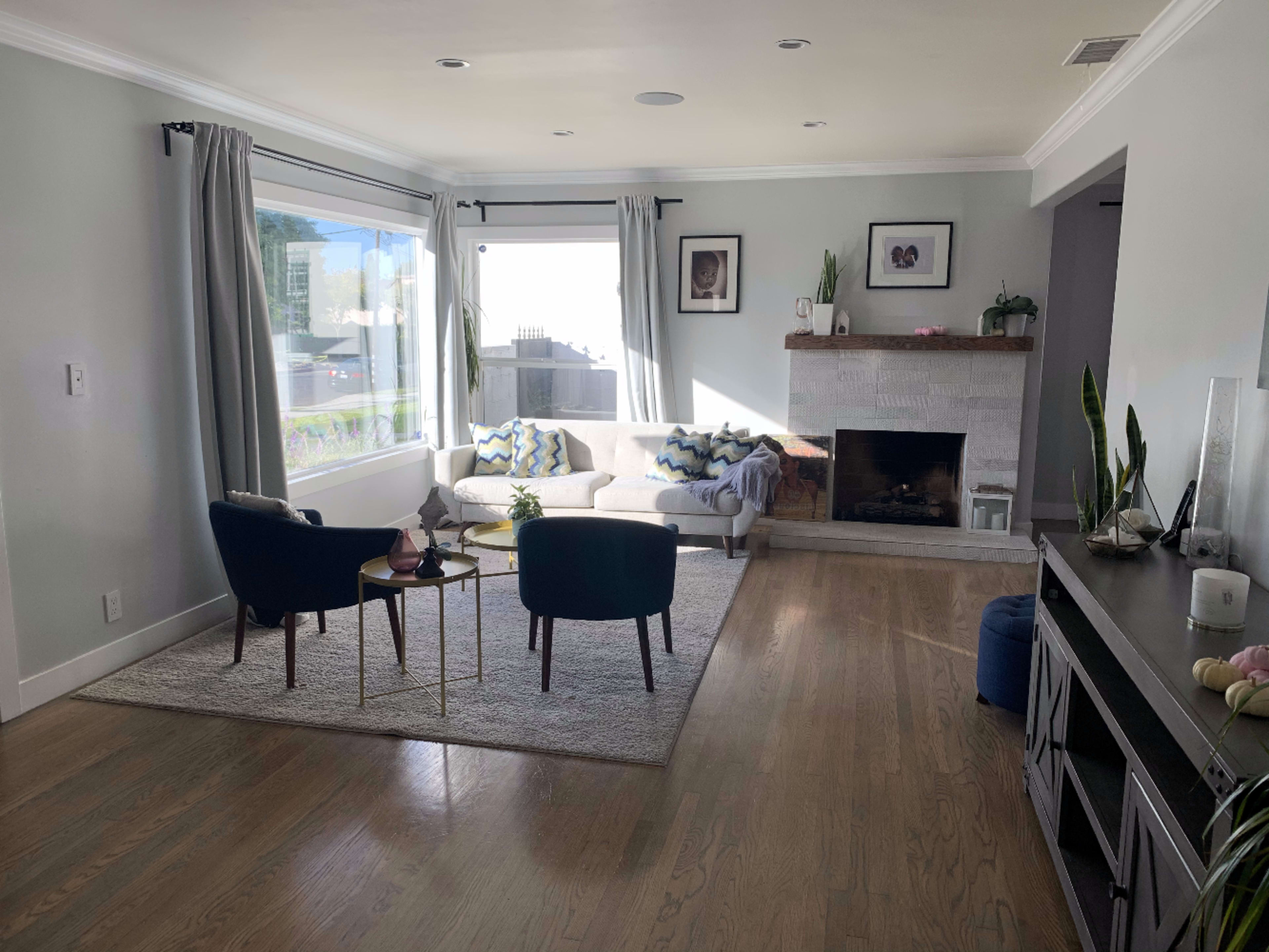A living room features a large window with sheer curtains, a light-colored sofa, two chairs, a small coffee table, and a stone fireplace.