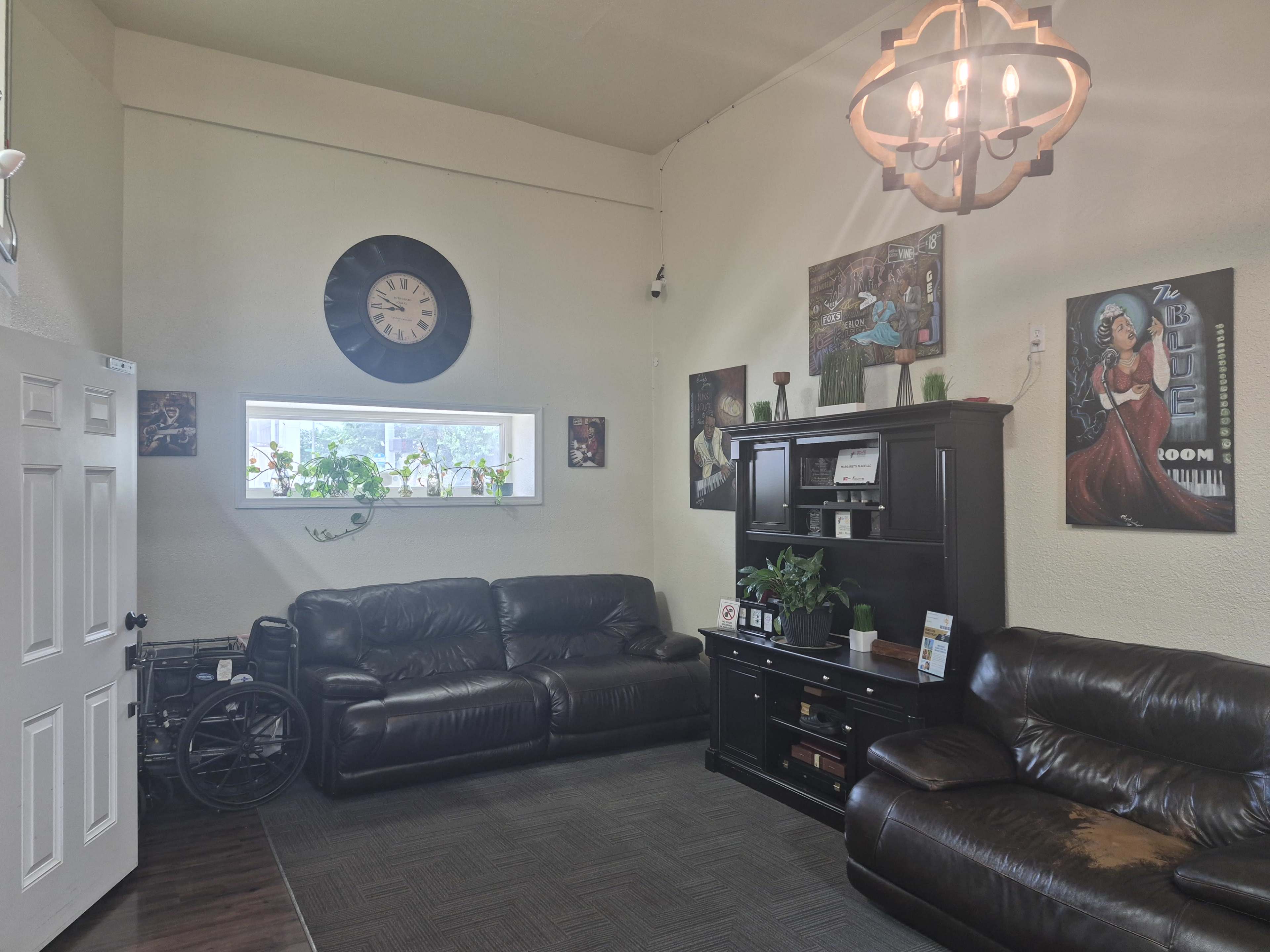 A cozy living room features two dark leather sofas, a black cabinet, framed artwork on the walls, and a large clock above a window.