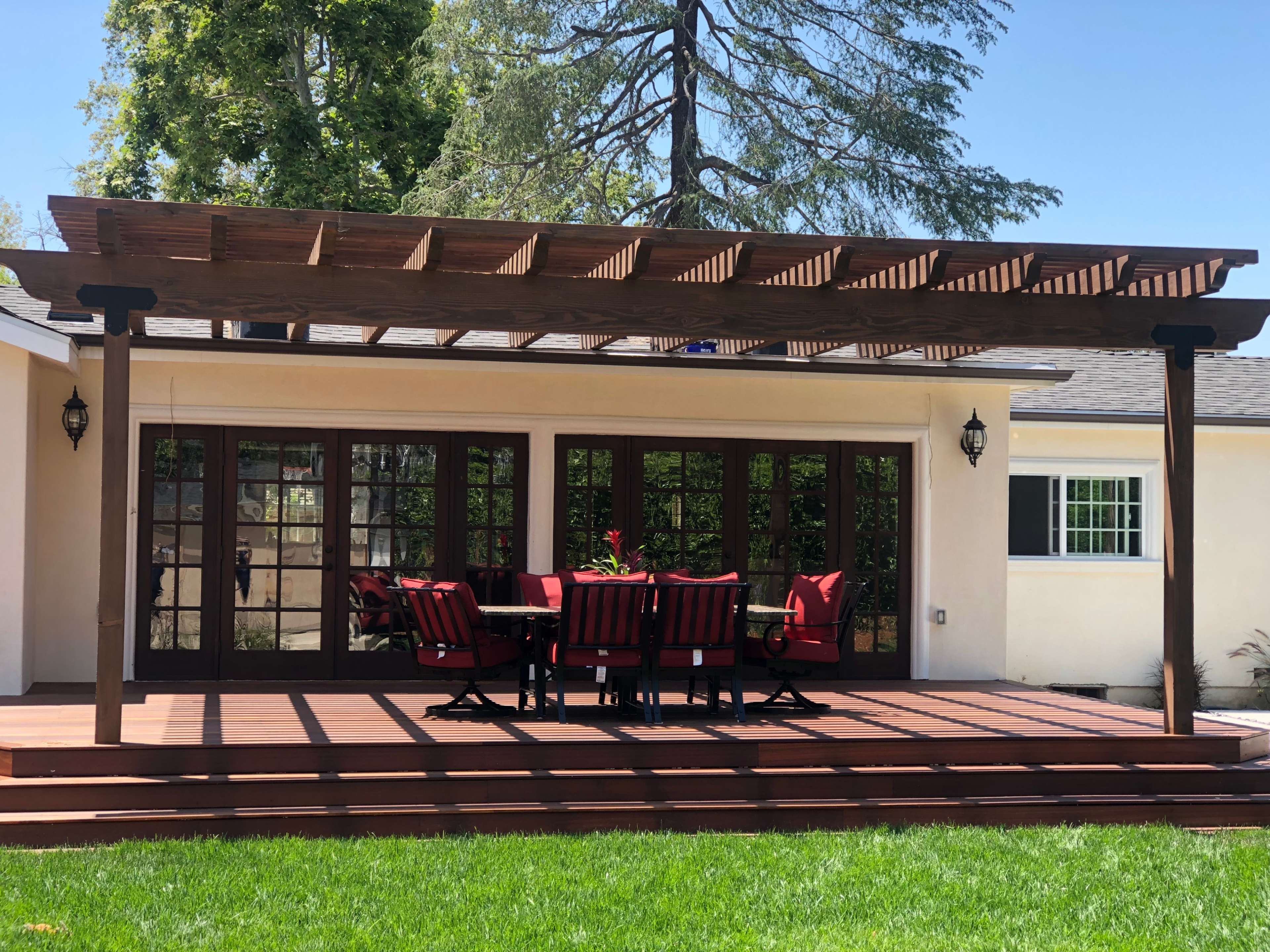 A wooden pergola stands over a dining table with red chairs on a deck next to a house surrounded by grass and trees.