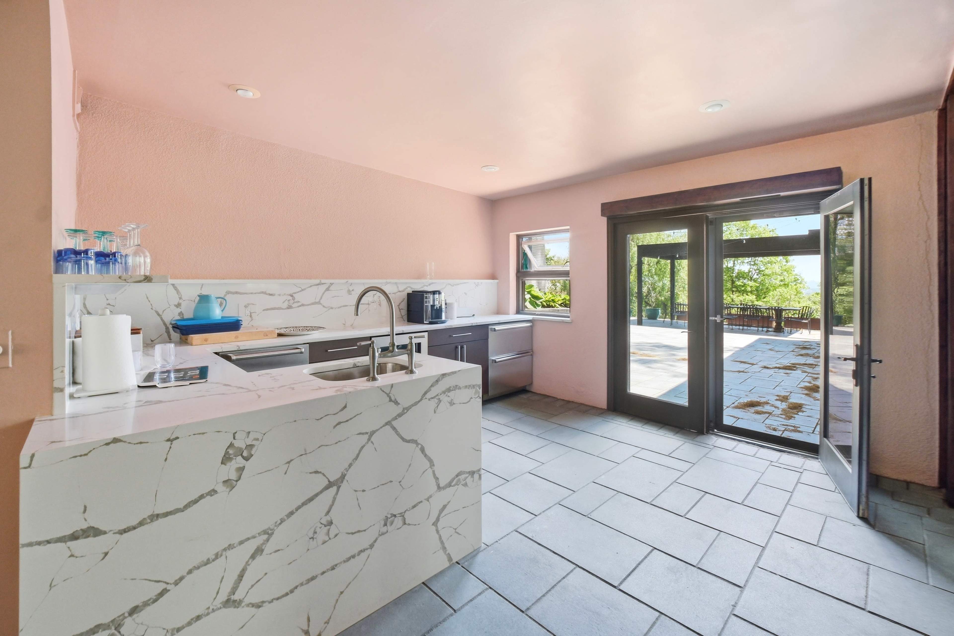 The kitchen features a pink wall, a marble countertop with a sink, modern appliances, and a doorway leading to an outdoor patio.