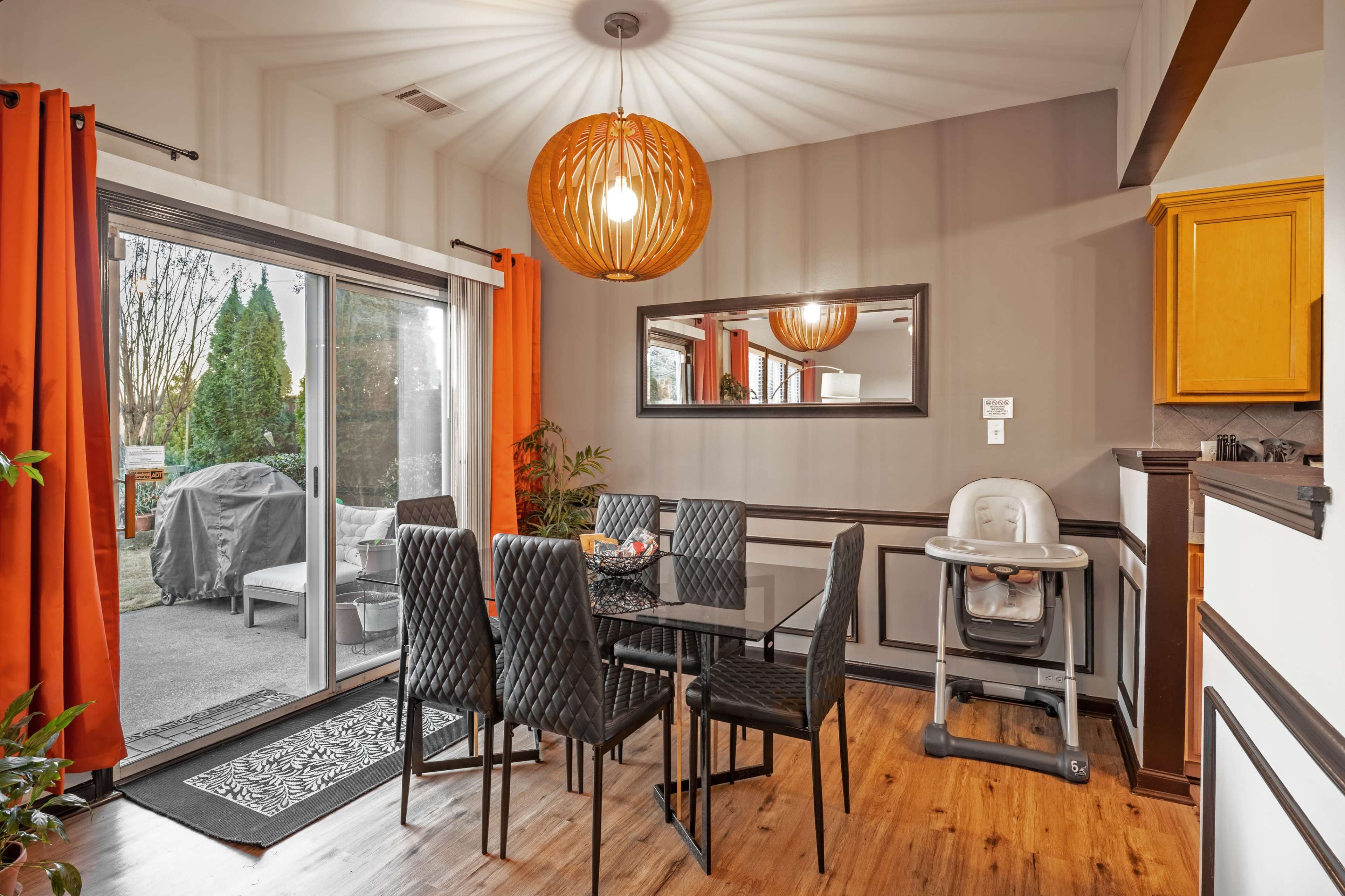 A dining area features a glass table surrounded by black chairs, with an orange pendant light overhead and a sliding door leading to an outdoor space.