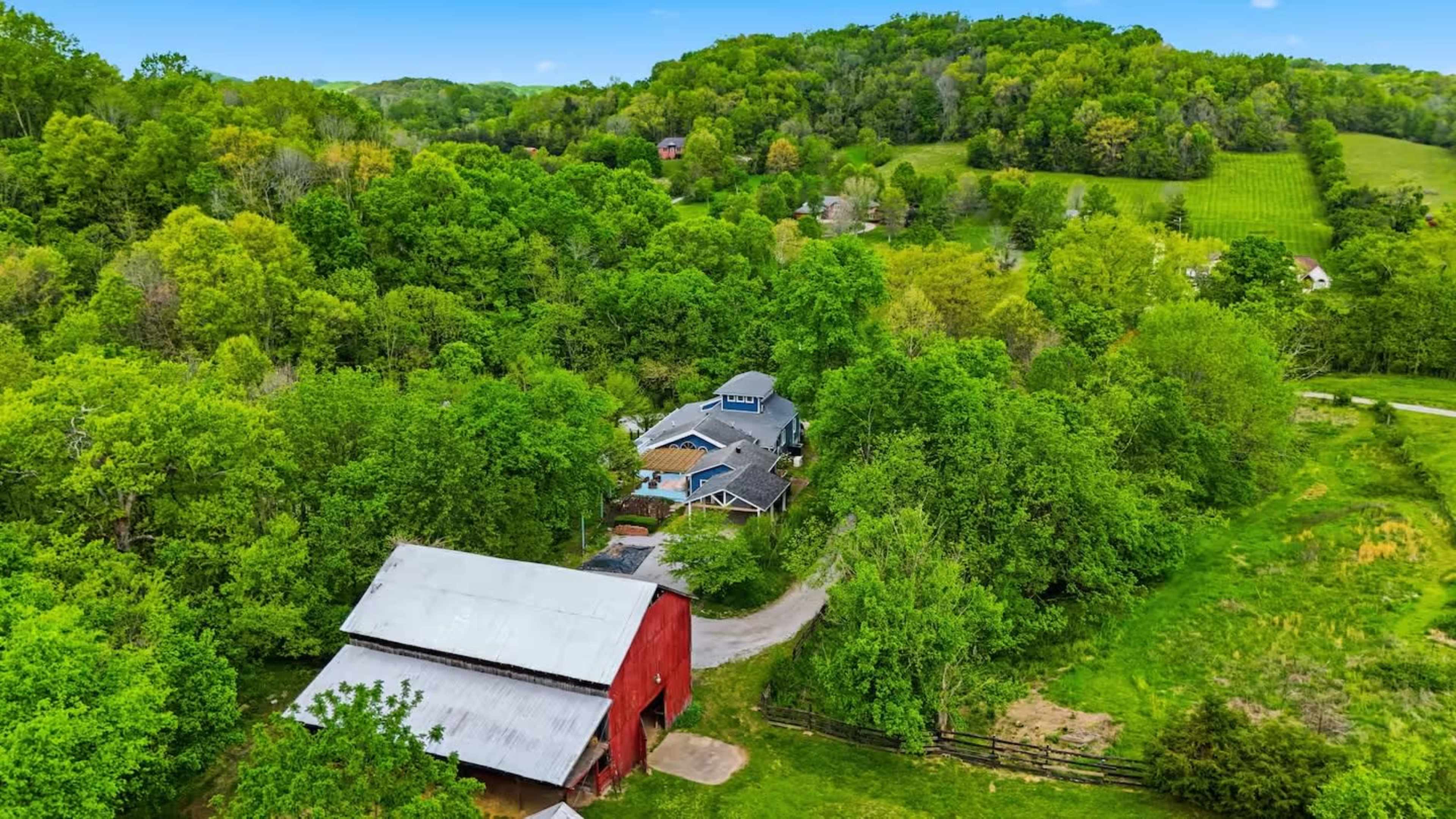 A red barn is situated near a house surrounded by lush green trees and rolling hills in a rural landscape.