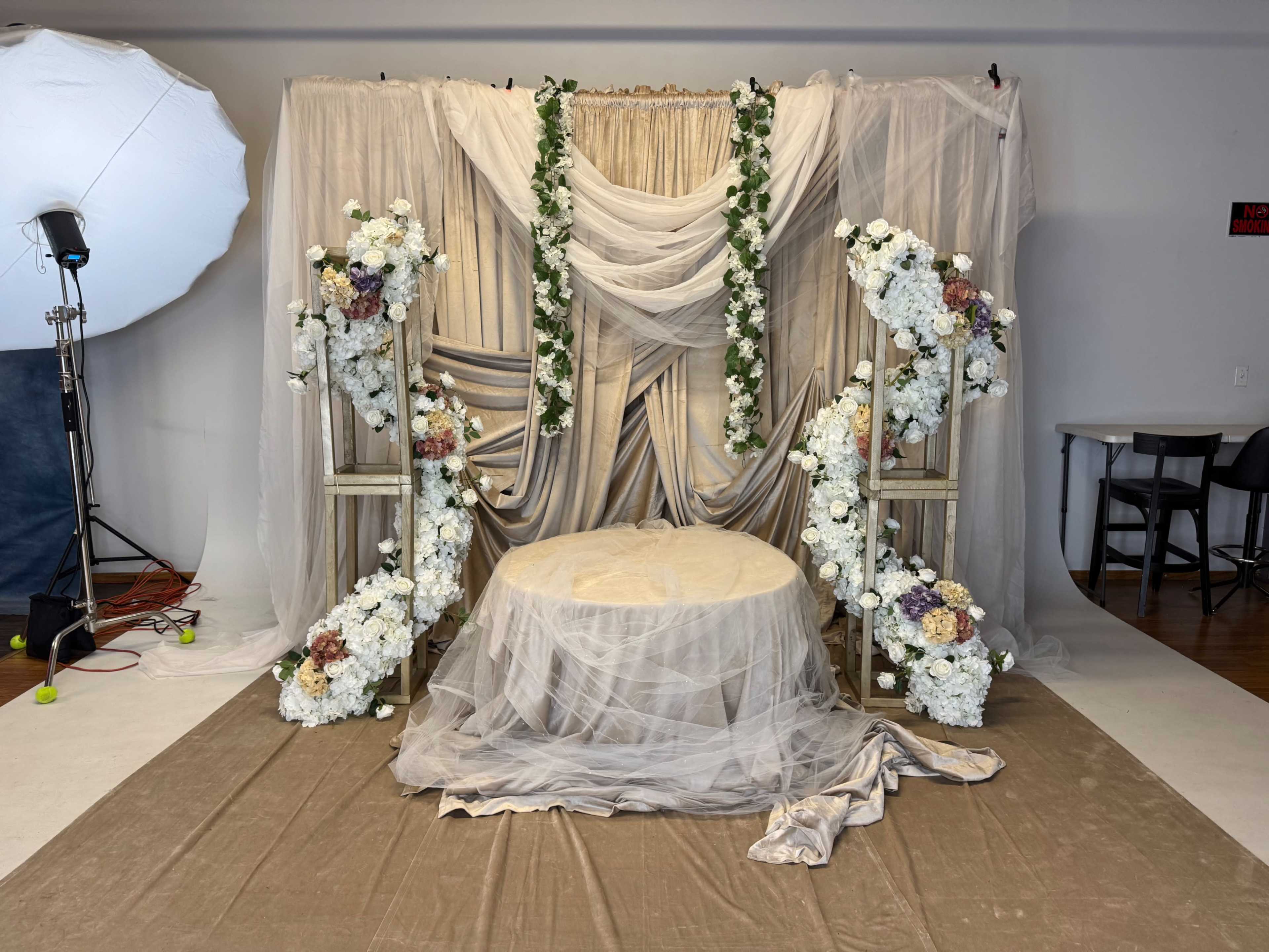 The image shows a decorative backdrop featuring draped fabric and floral arrangements, set up with a circular table in front.