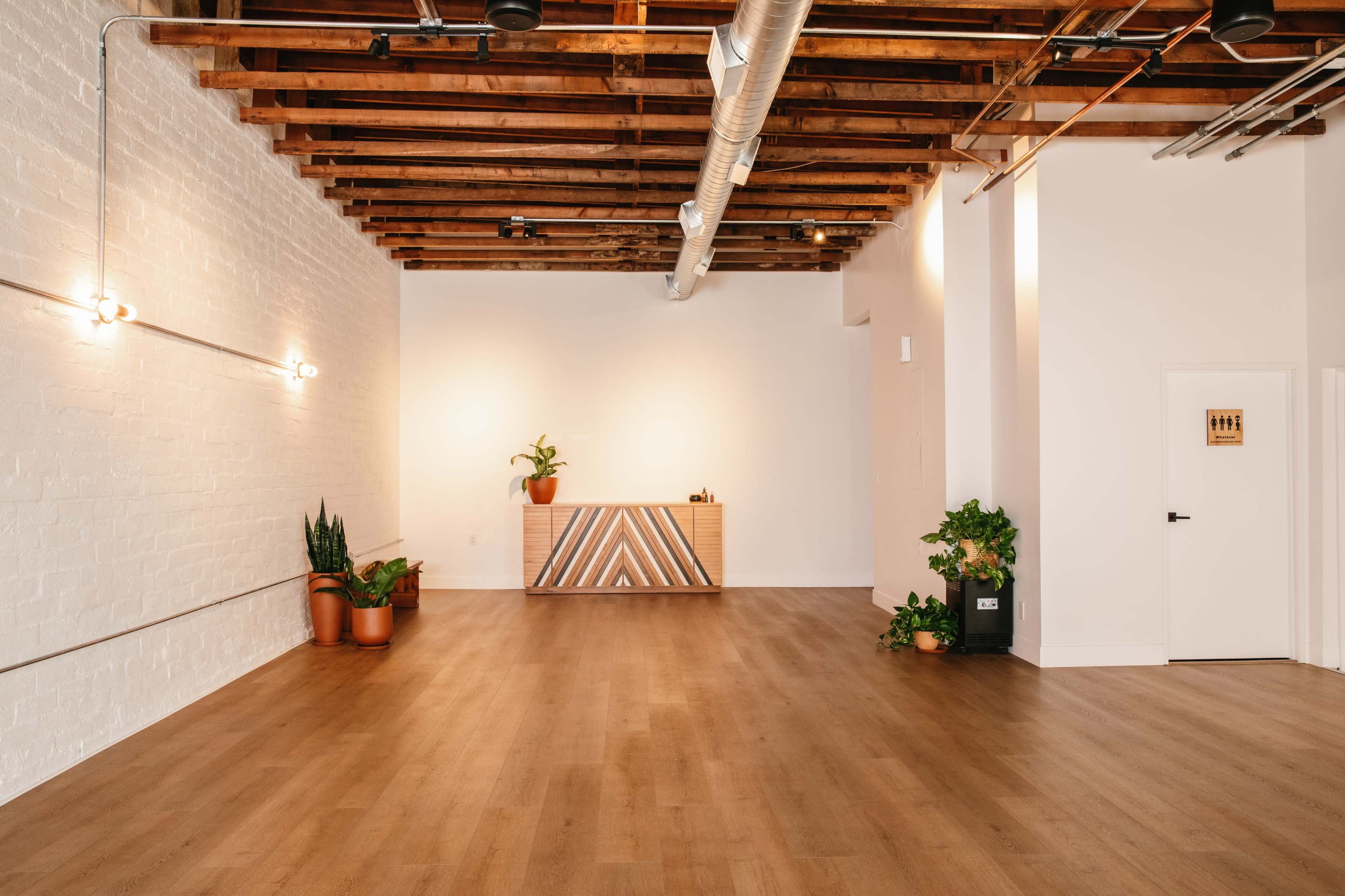 The image shows a spacious, empty room with wooden flooring, exposed beams, and a reception desk against a white wall.