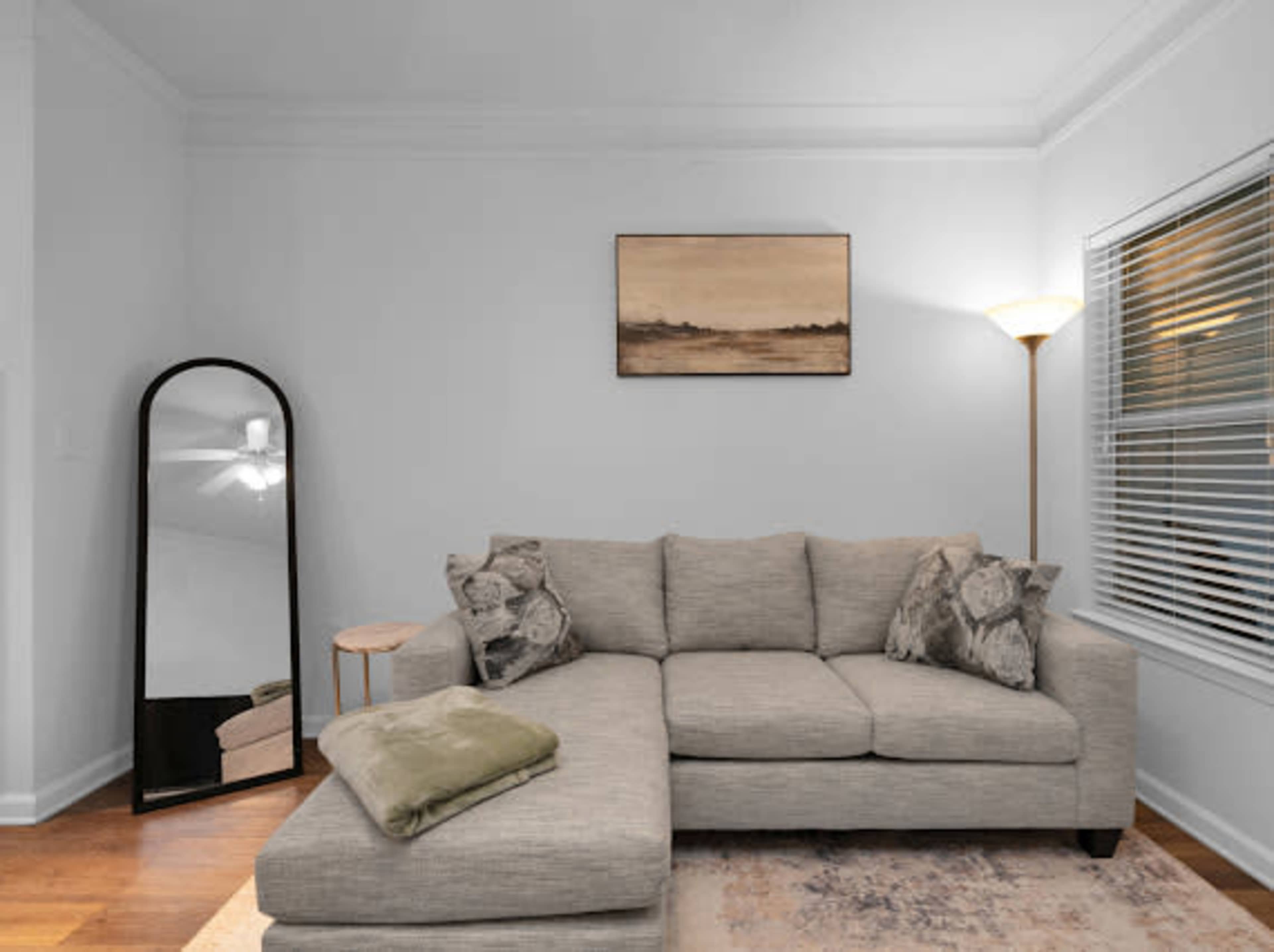 The image depicts a modern living room with a gray sectional sofa, a decorative throw blanket, a floor lamp, a side table, and a tall mirror against the wall.