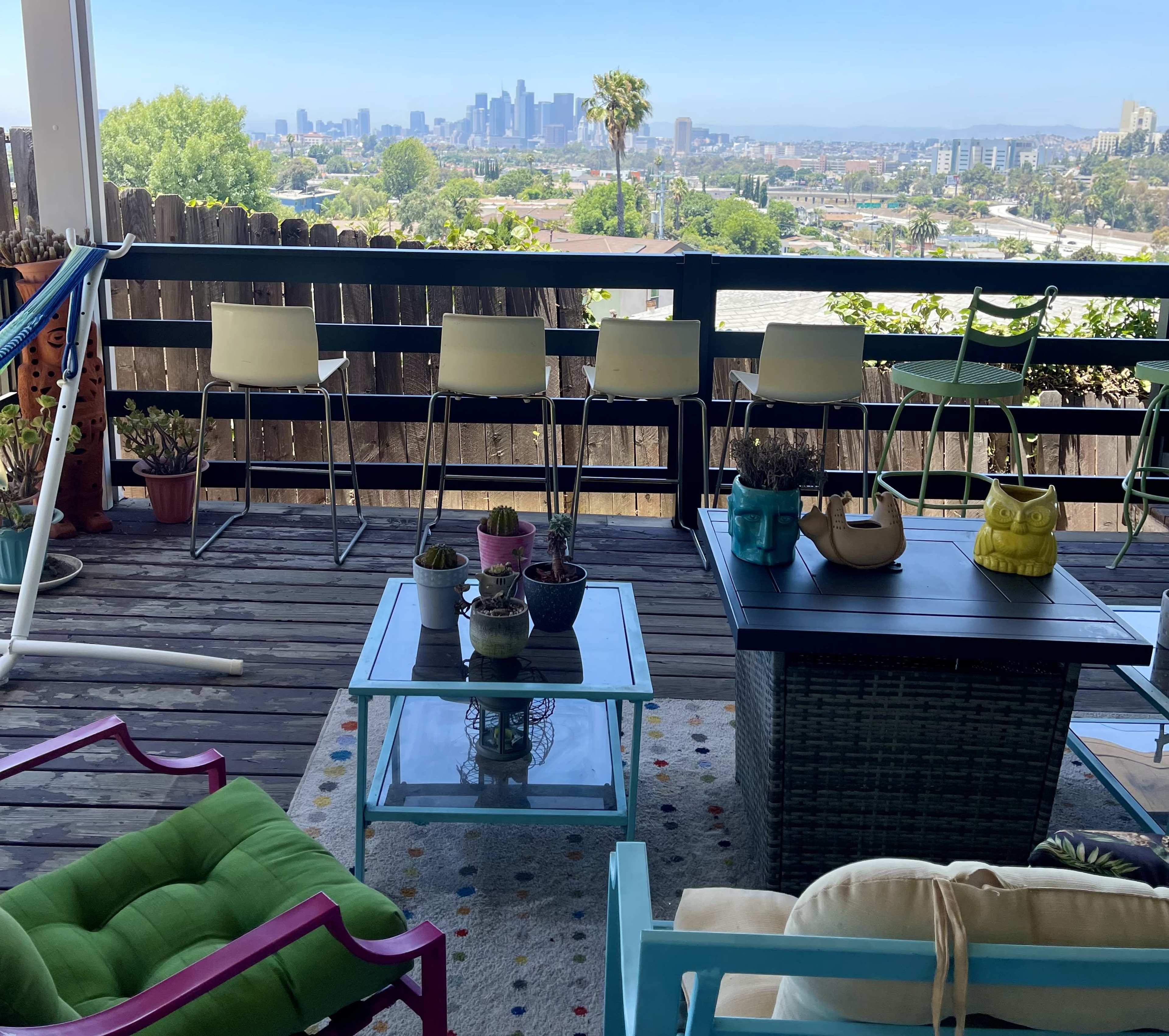 A shaded balcony features colorful outdoor furniture and planters, overlooking a city skyline with distant hills.