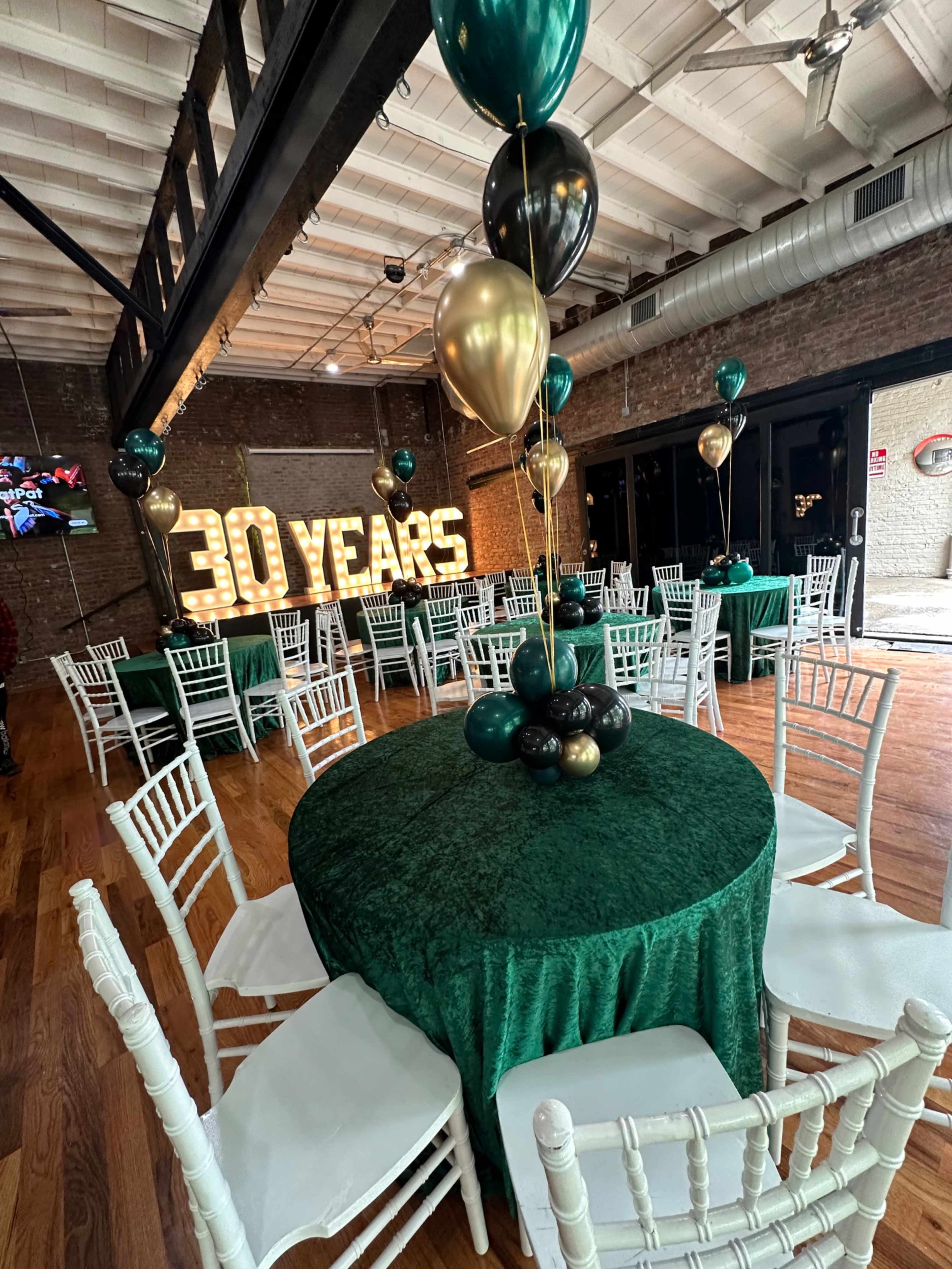 The image shows a festive event space with round tables covered in green cloth, white chairs, and balloons in black and gold, alongside a large illuminated "30 YEARS" sign.