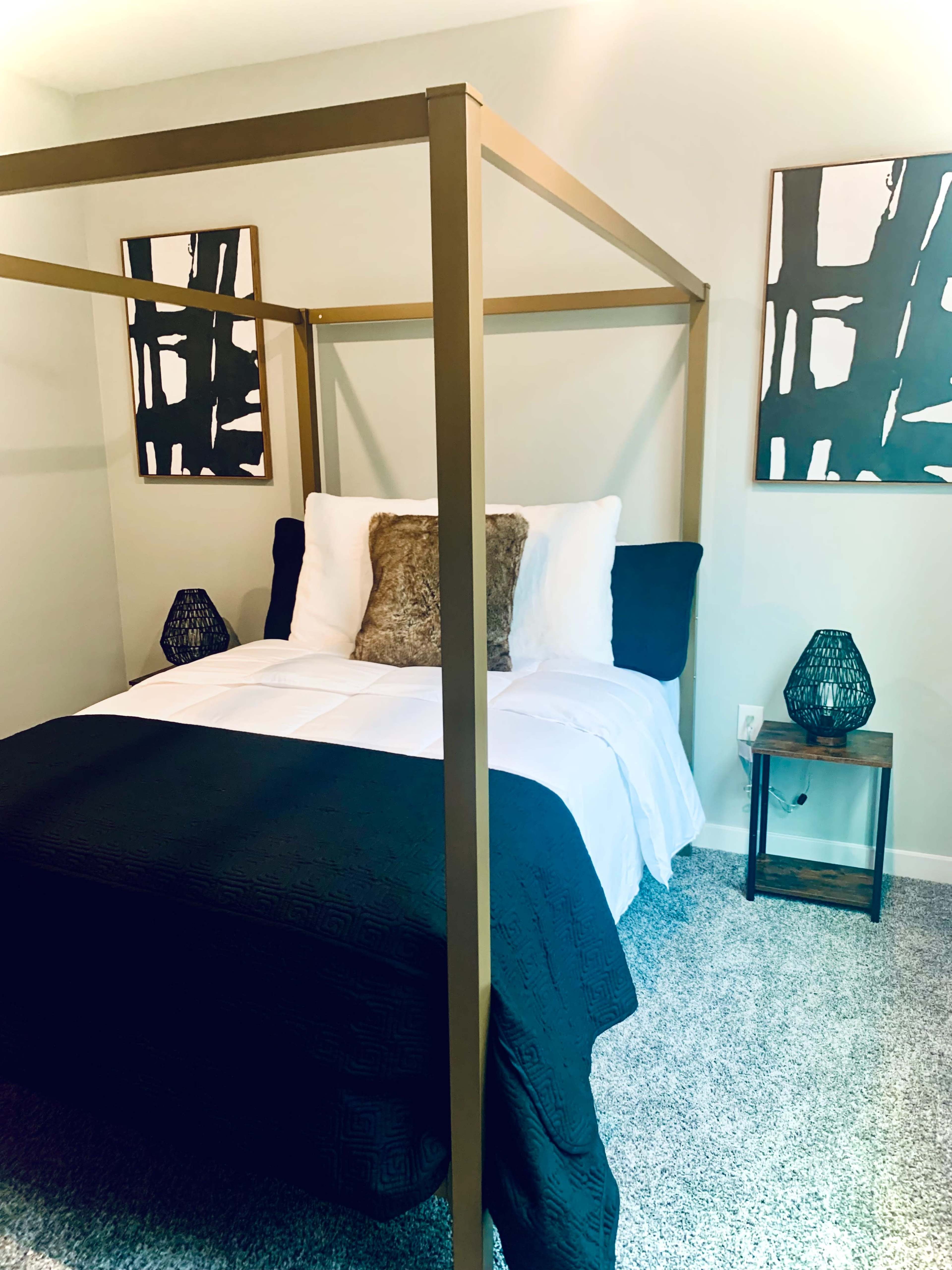 A canopy bed with a dark bedspread and decorative pillows, flanked by two patterned wall art pieces and small lamps on either side.