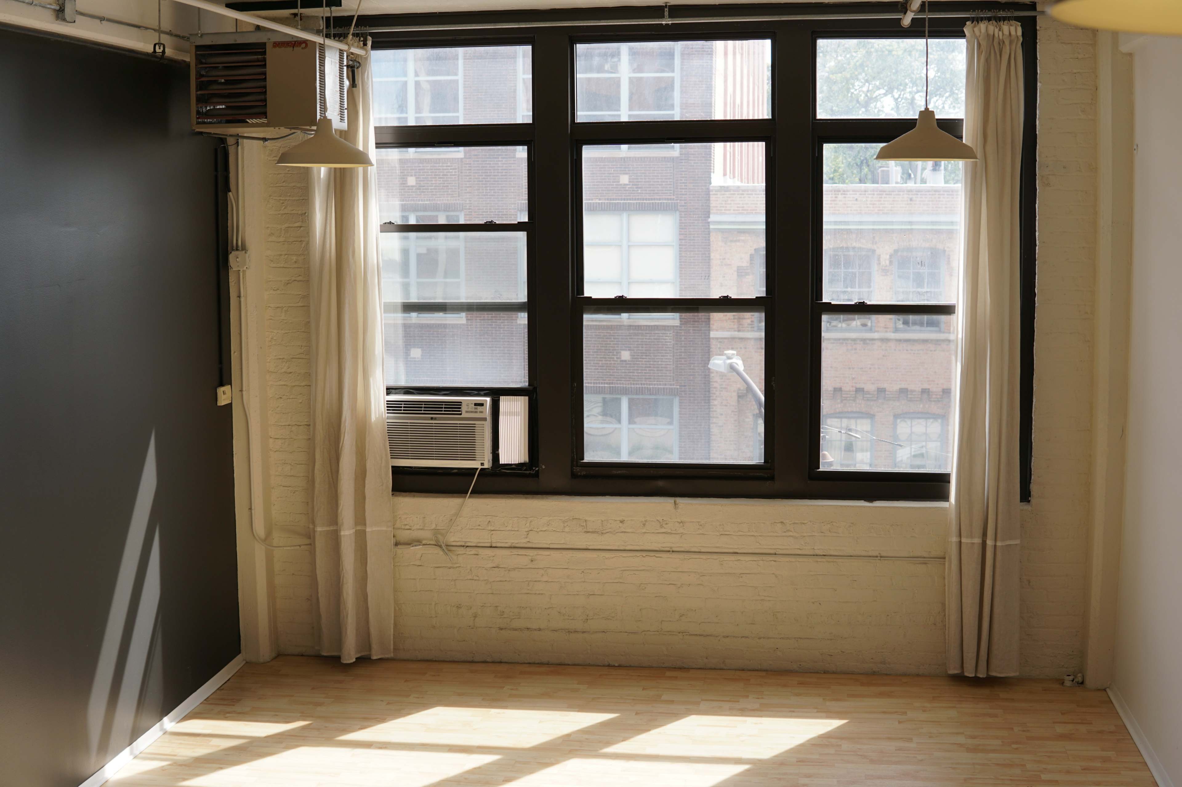 A sunlit, empty room with large windows featuring an air conditioning unit and light curtains.