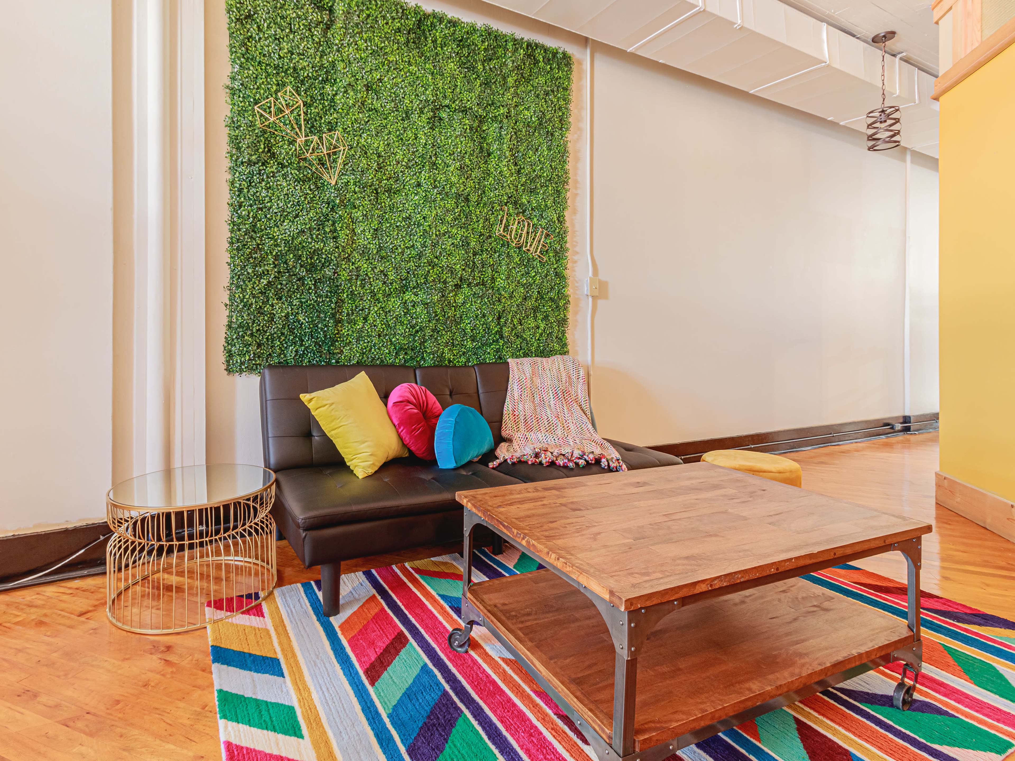 The image shows a modern lounge area with a black sofa, colorful cushions, a wooden coffee table, and a vibrant striped rug, adjacent to a green wall of artificial plants.