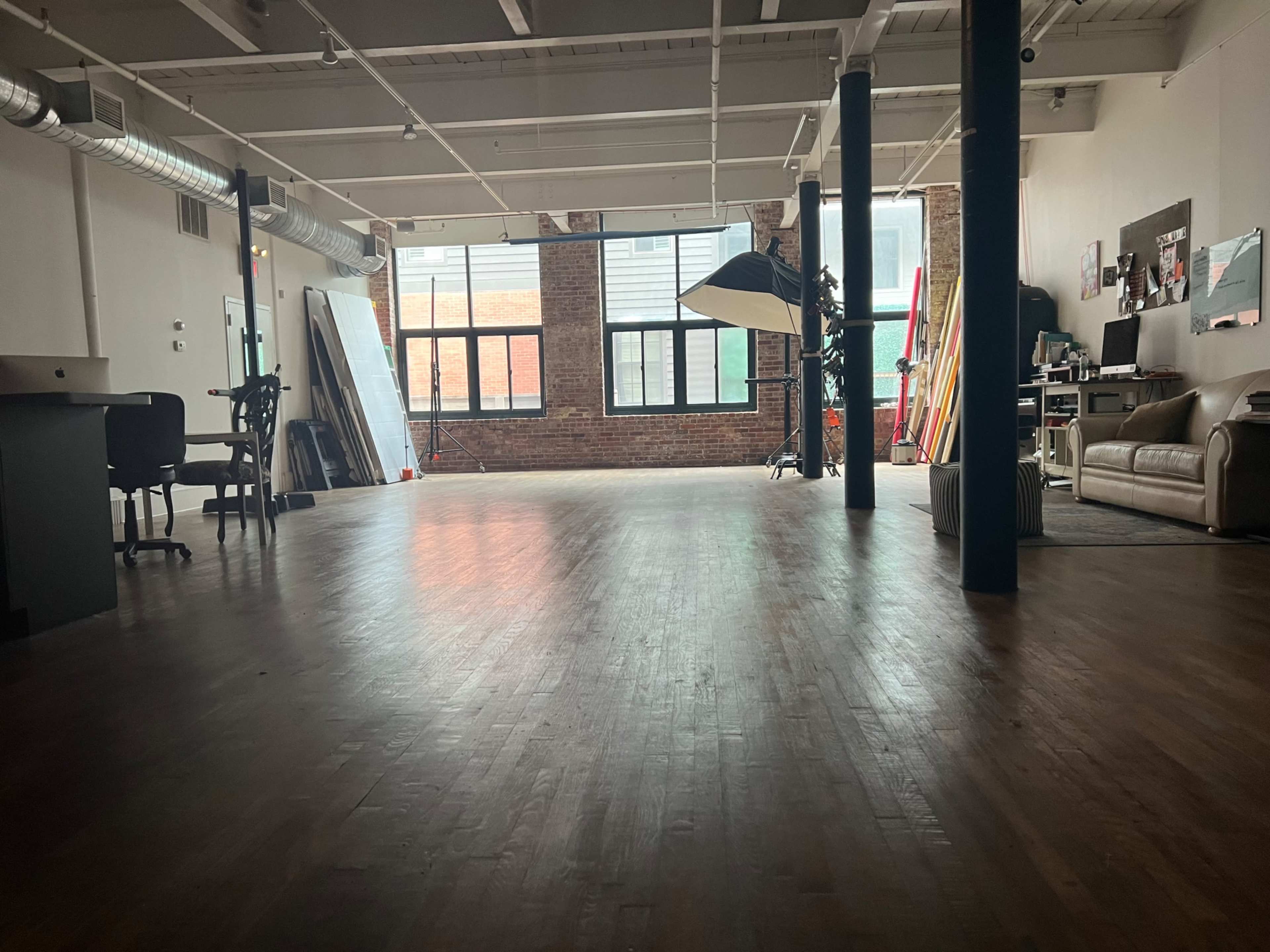 A spacious, industrial-style room features hardwood flooring, large windows, and various furnishings including a desk and a couch.