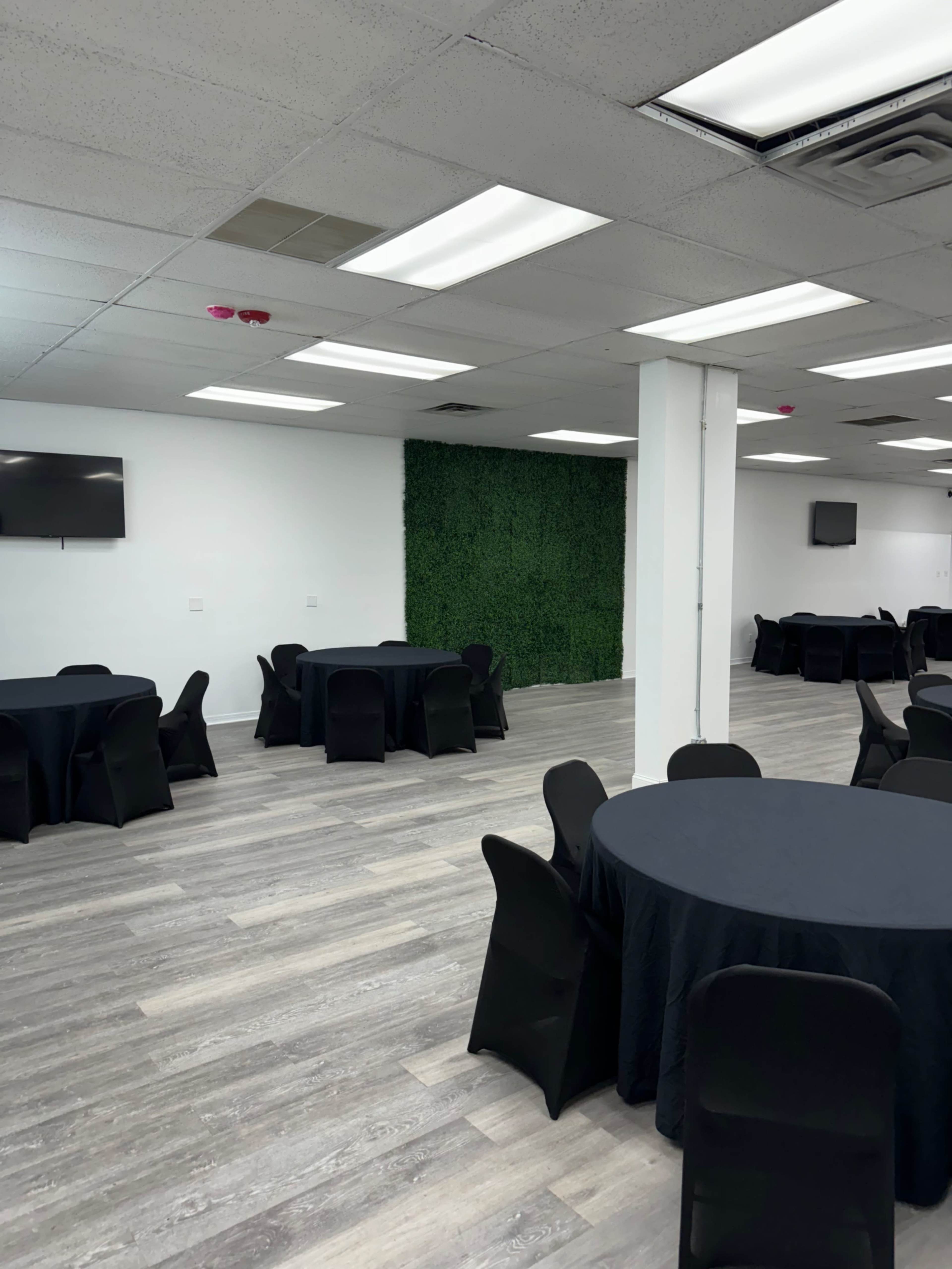 A spacious room with several round black tables covered with black tablecloths and a green wall of plants at one end.