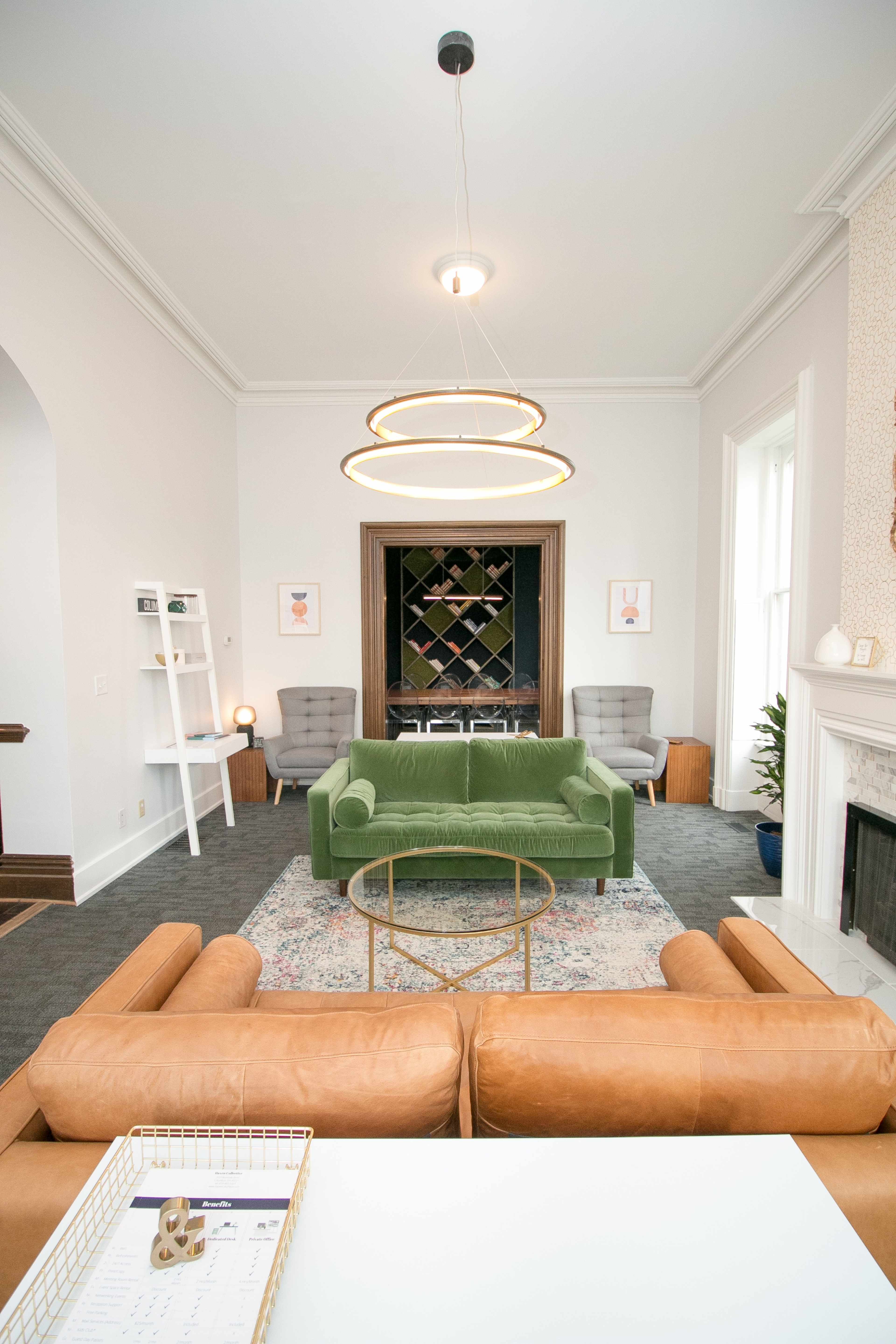 A modern living room with a green velvet sofa, two gray armchairs, a circular chandelier, and a wine rack in the background.