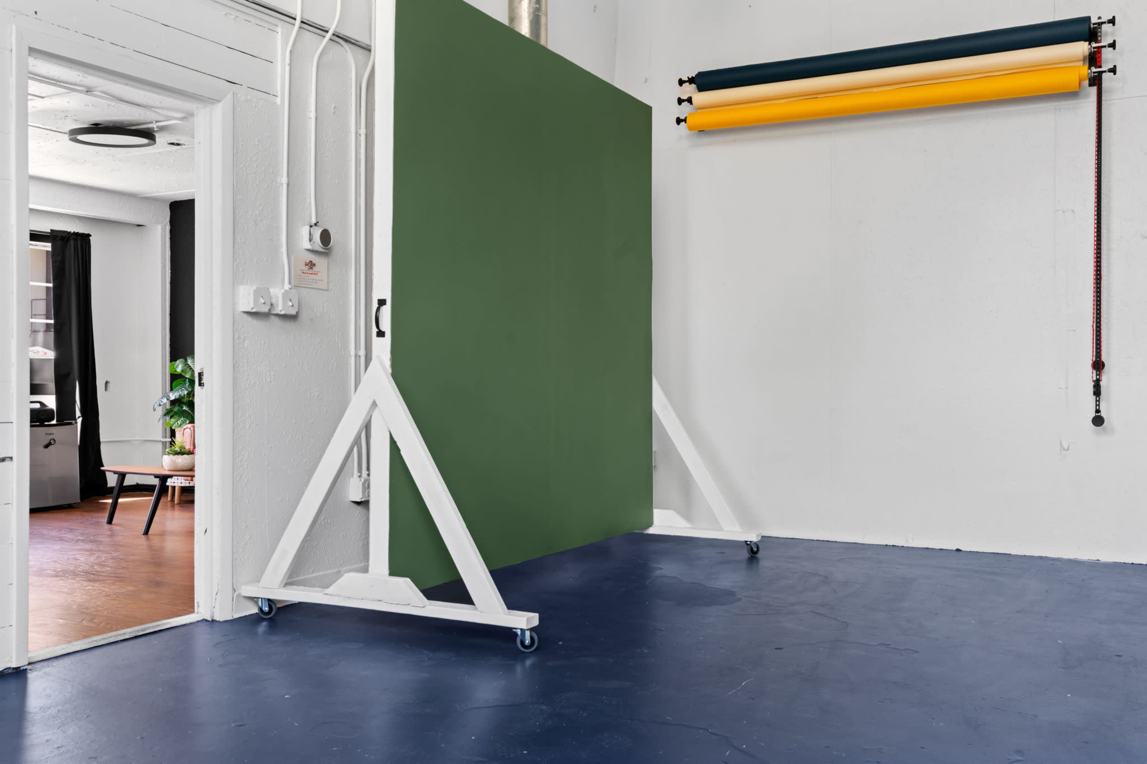 A studio space featuring a green backdrop on wheels, with rolls of colored paper hanging on a wall and a doorway leading to another room.