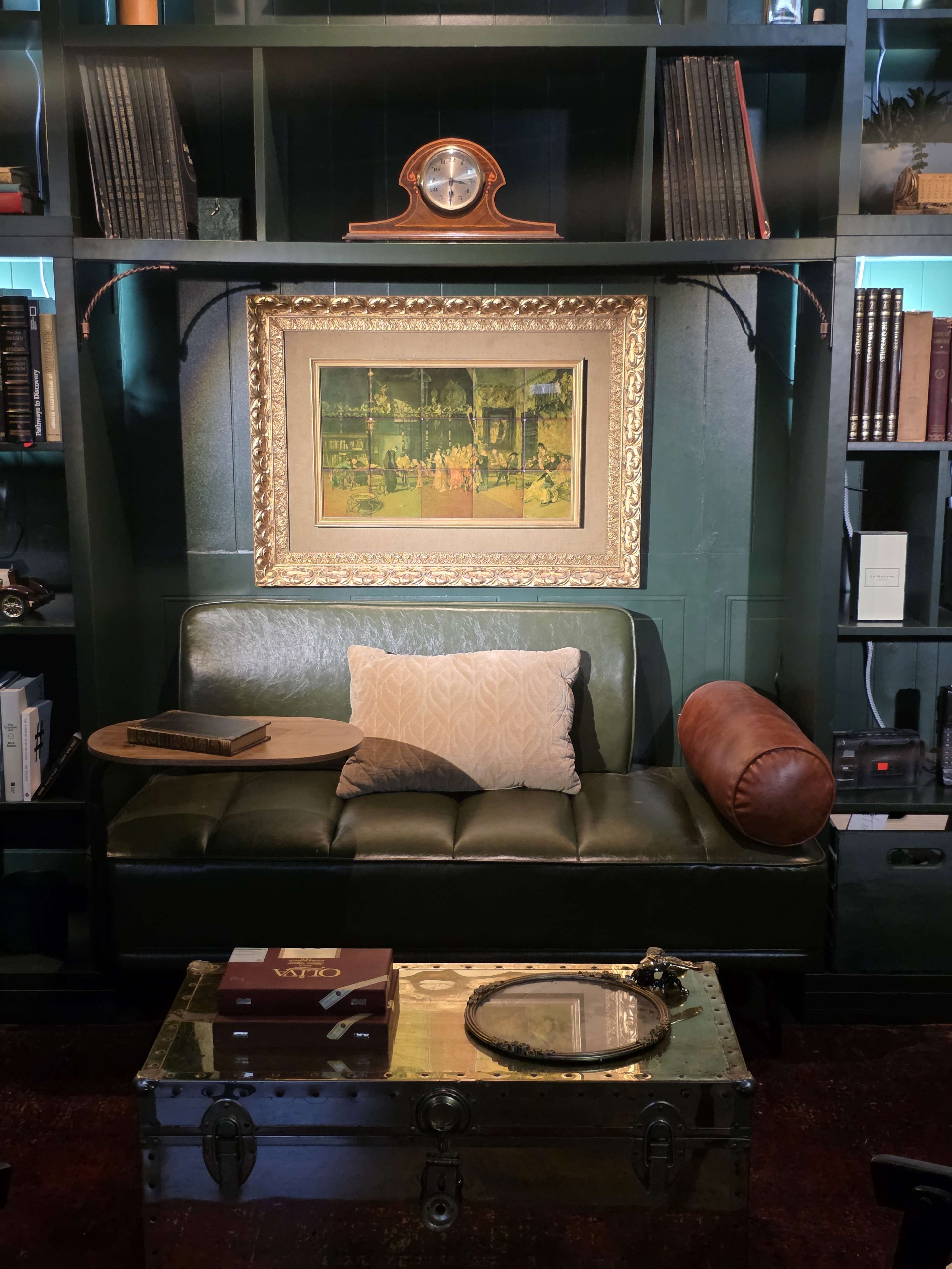 A green leather sofa is positioned in front of a bookshelf, featuring a framed painting above it and a small side table beside the couch.