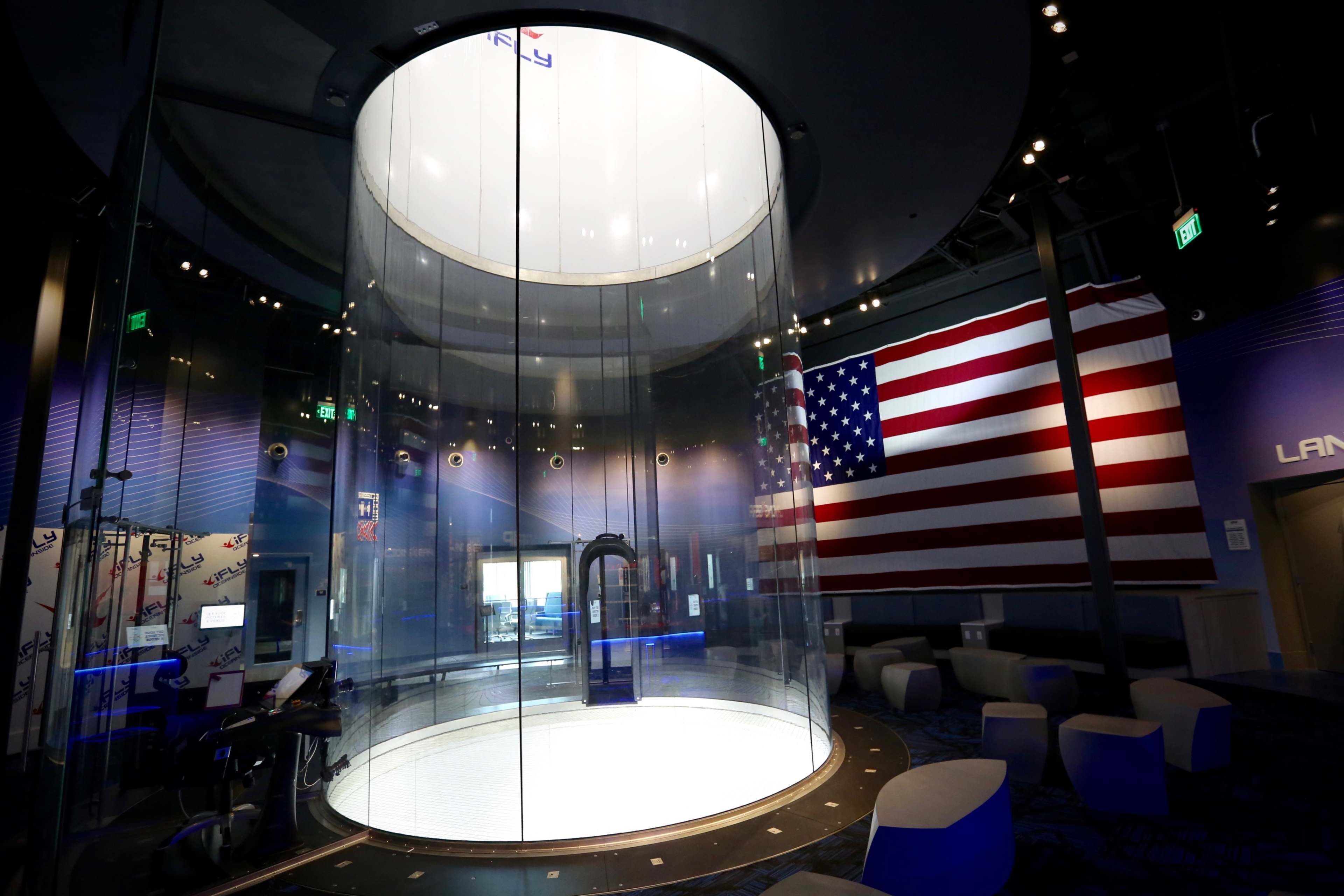 A spacious indoor skydiving facility featuring a large transparent flight chamber and a prominent American flag on the wall.
