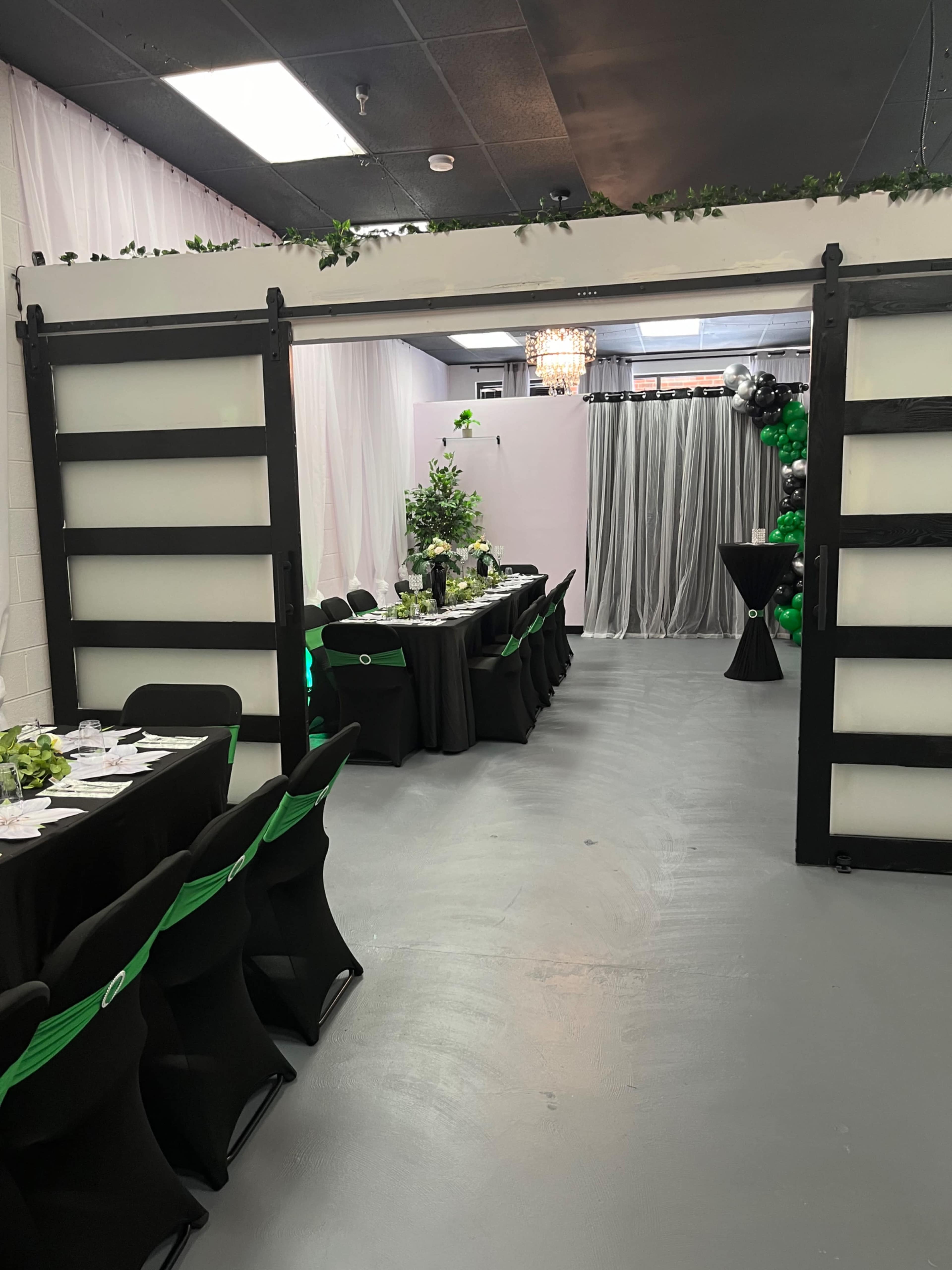 The image shows a decorated event space featuring long tables with green and black table settings, flanked by a lush plant centerpiece, and a backdrop of balloons and draped fabric.