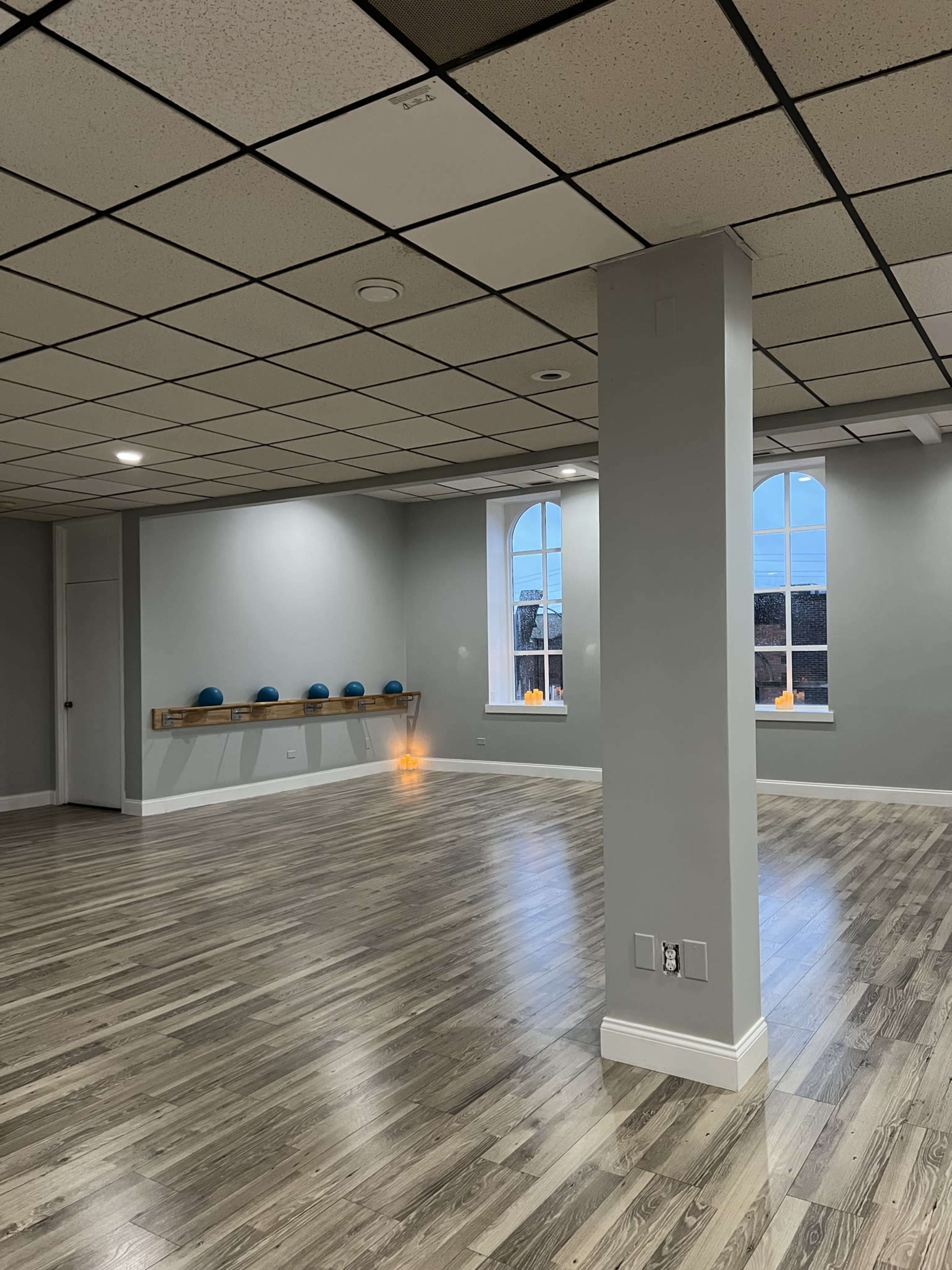 A spacious, empty studio with wooden flooring, large windows, and a wall-mounted shelf holding exercise equipment.