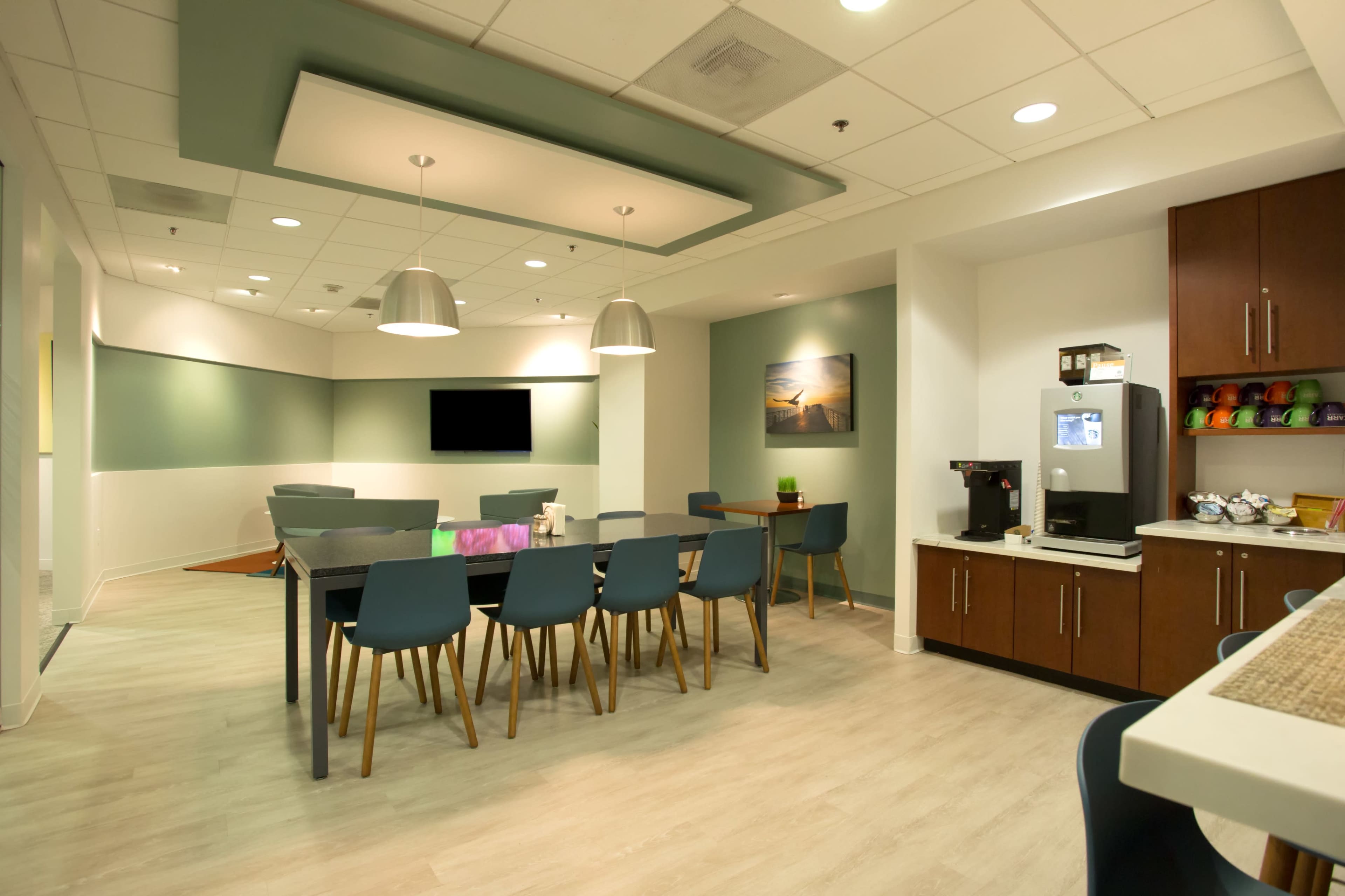 A modern conference room features a long table with blue chairs, lighting fixtures above, and a coffee station against the wall.