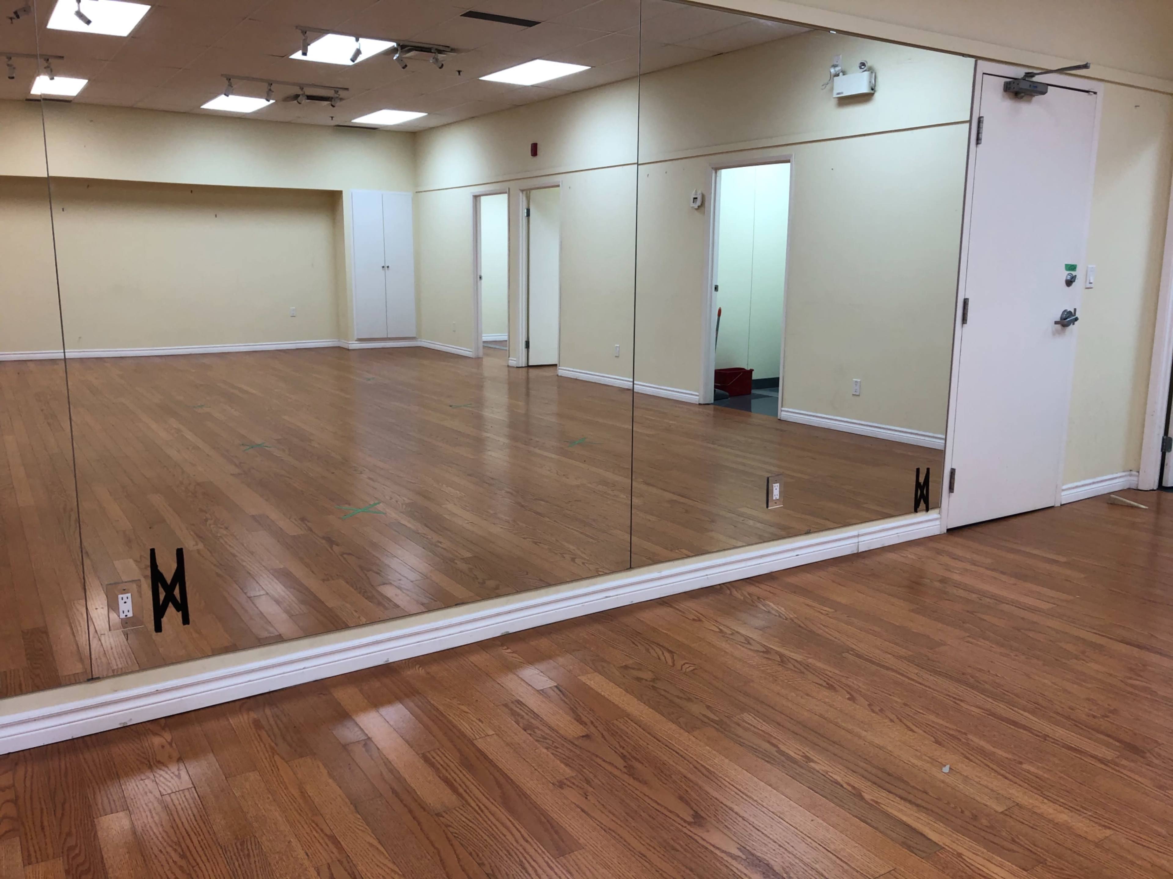 The image shows an empty room with wooden flooring, a large mirror along one wall, and a white door on the opposite side.