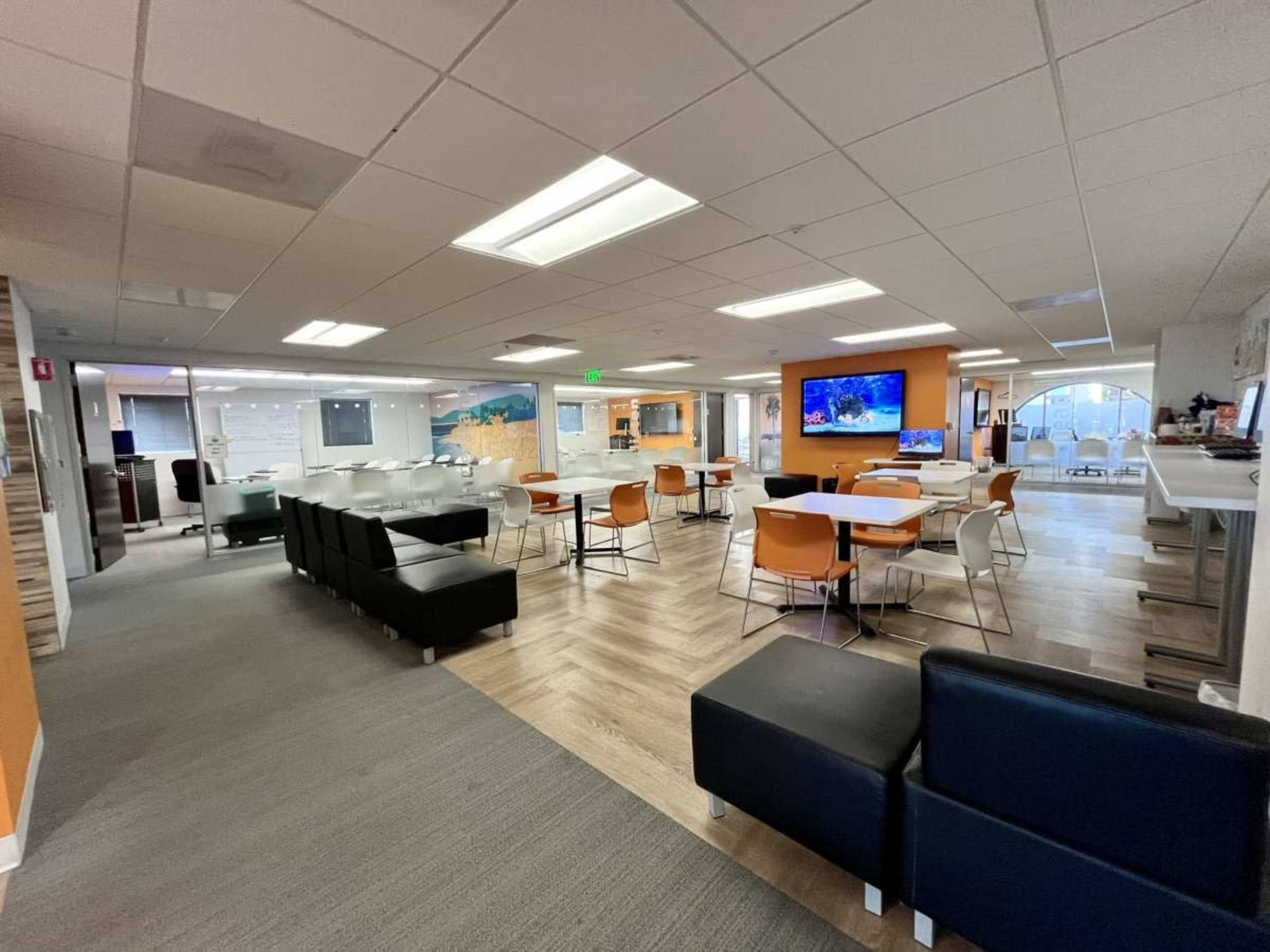 A modern office space featuring various seating arrangements, communal tables, and a large screen on the wall.