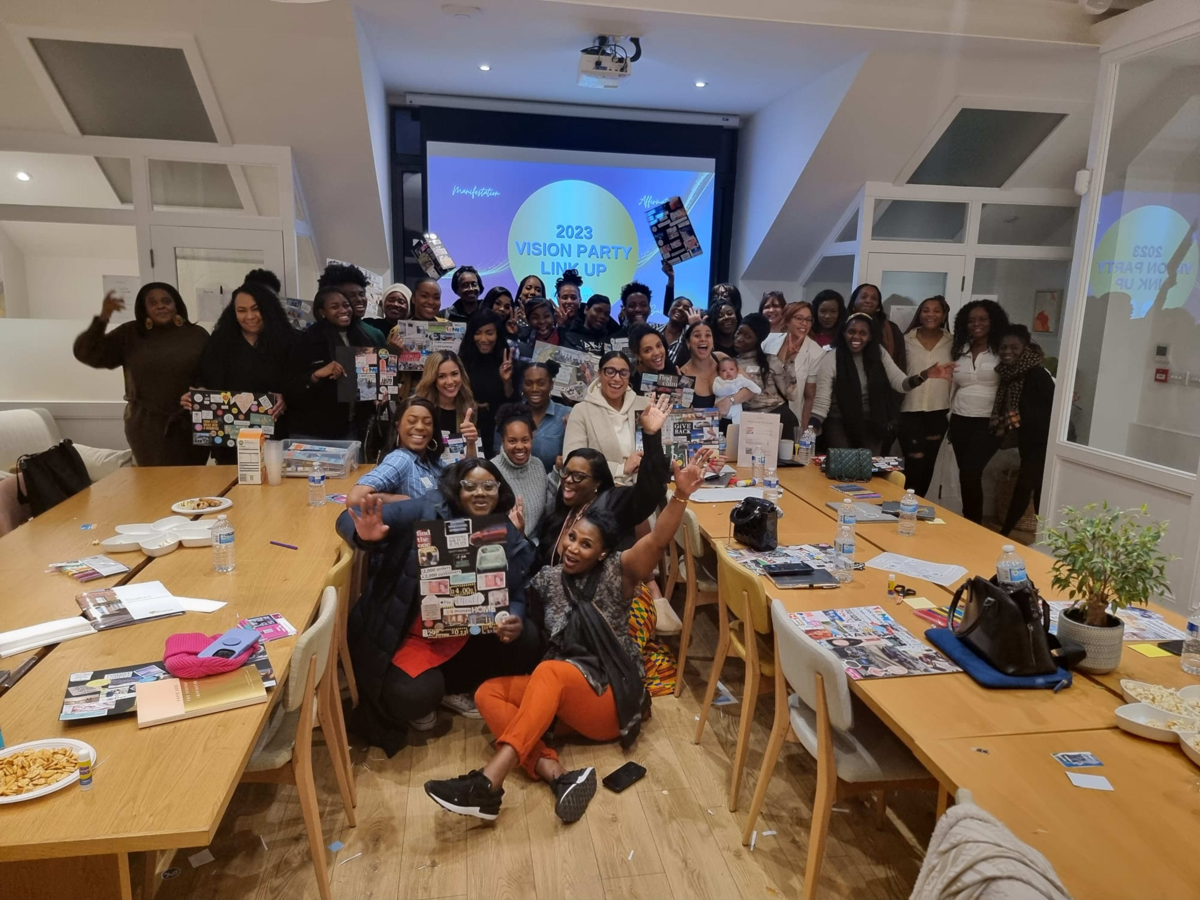 A large group of people gathers around a table in a well-lit room, holding up vision boards and smiling towards the camera during a vision party event.