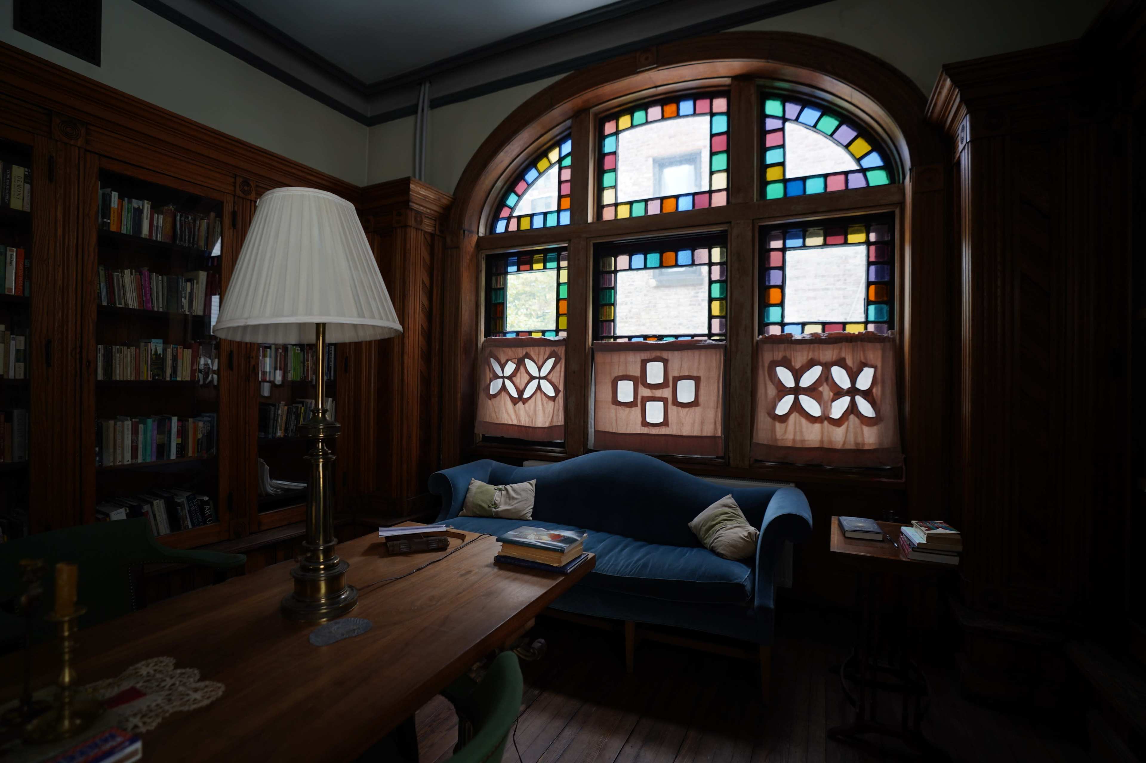 A well-furnished room features a wooden table and a blue sofa, with large stained glass windows illuminating the space.
