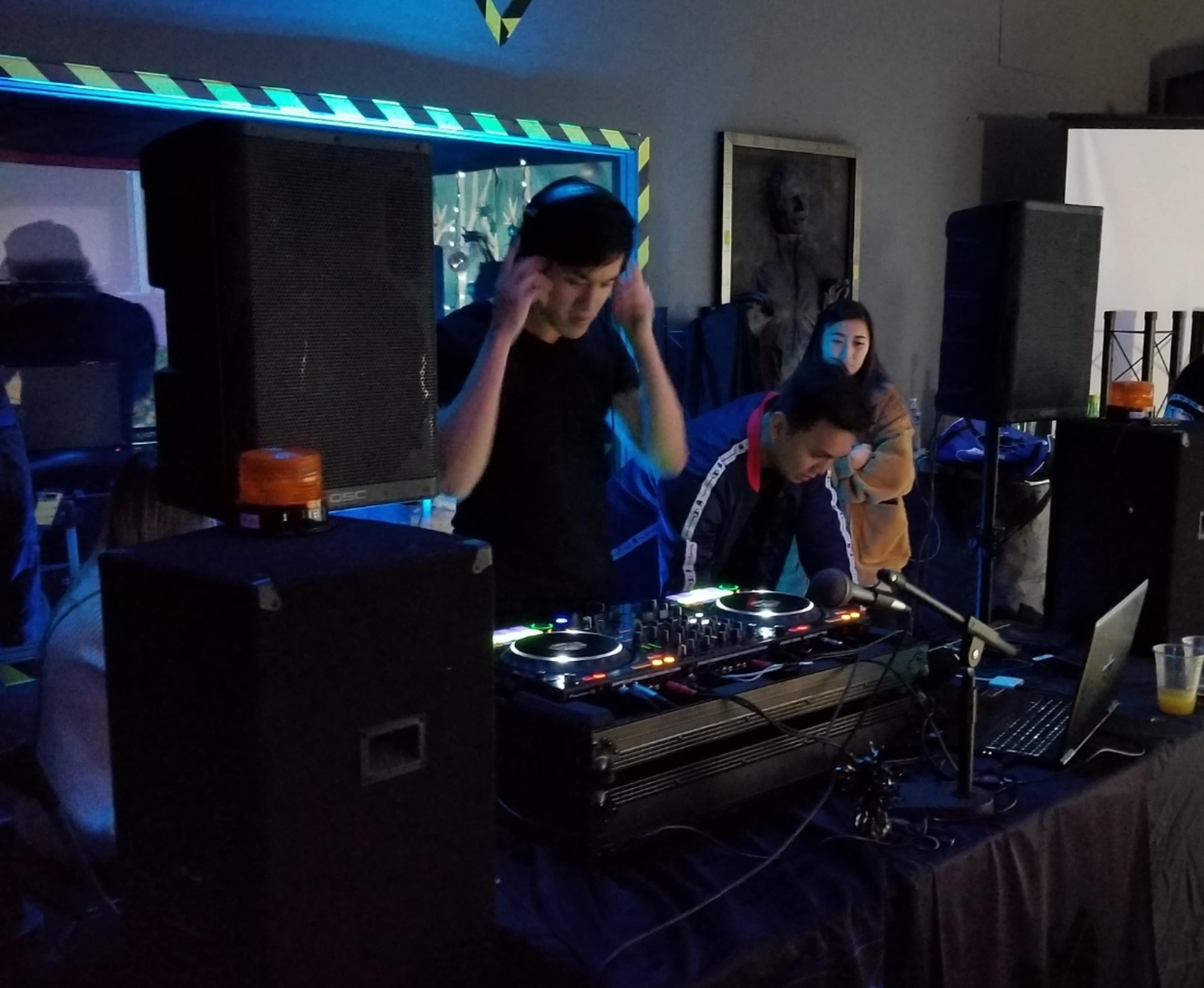 Two DJs are performing at a small venue with turntables, speakers, and a laptop, while a person watches in the background.