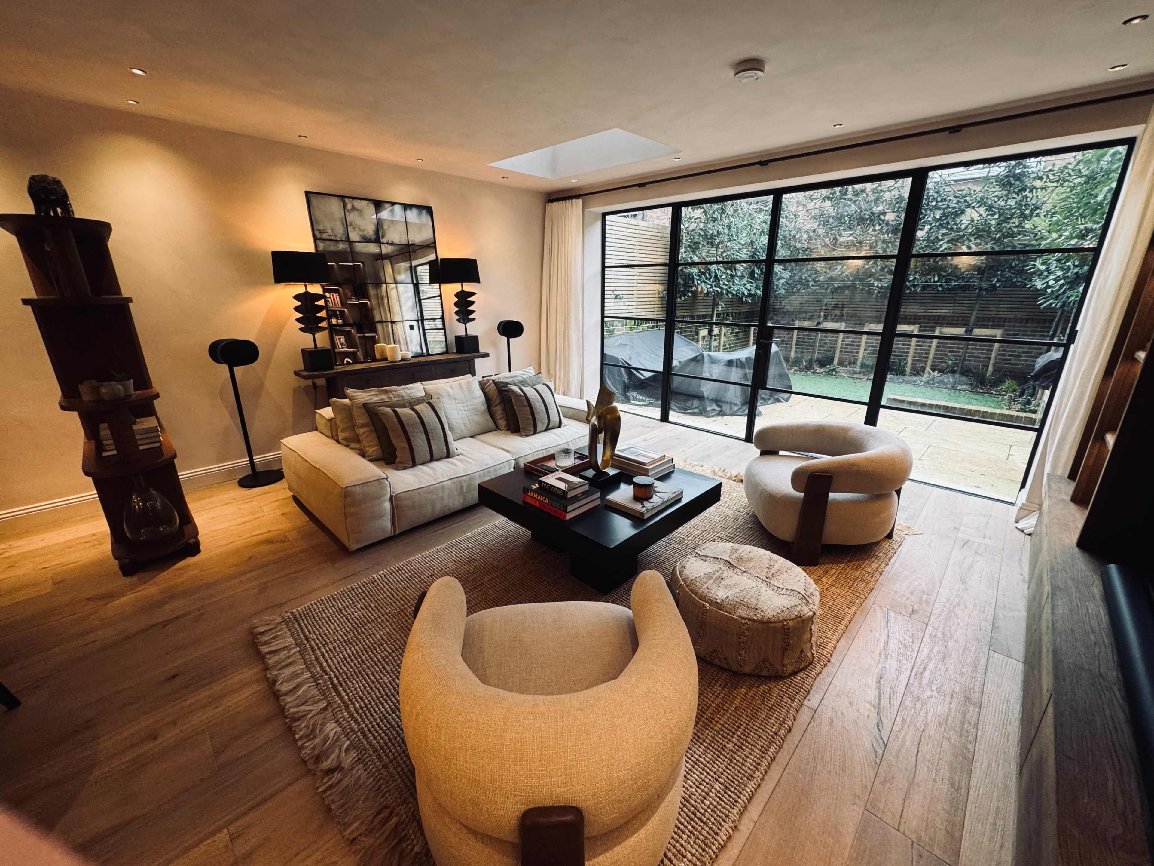 The image shows a modern living room featuring neutral-toned furniture, a large window overlooking a garden, and decorative elements on a coffee table.