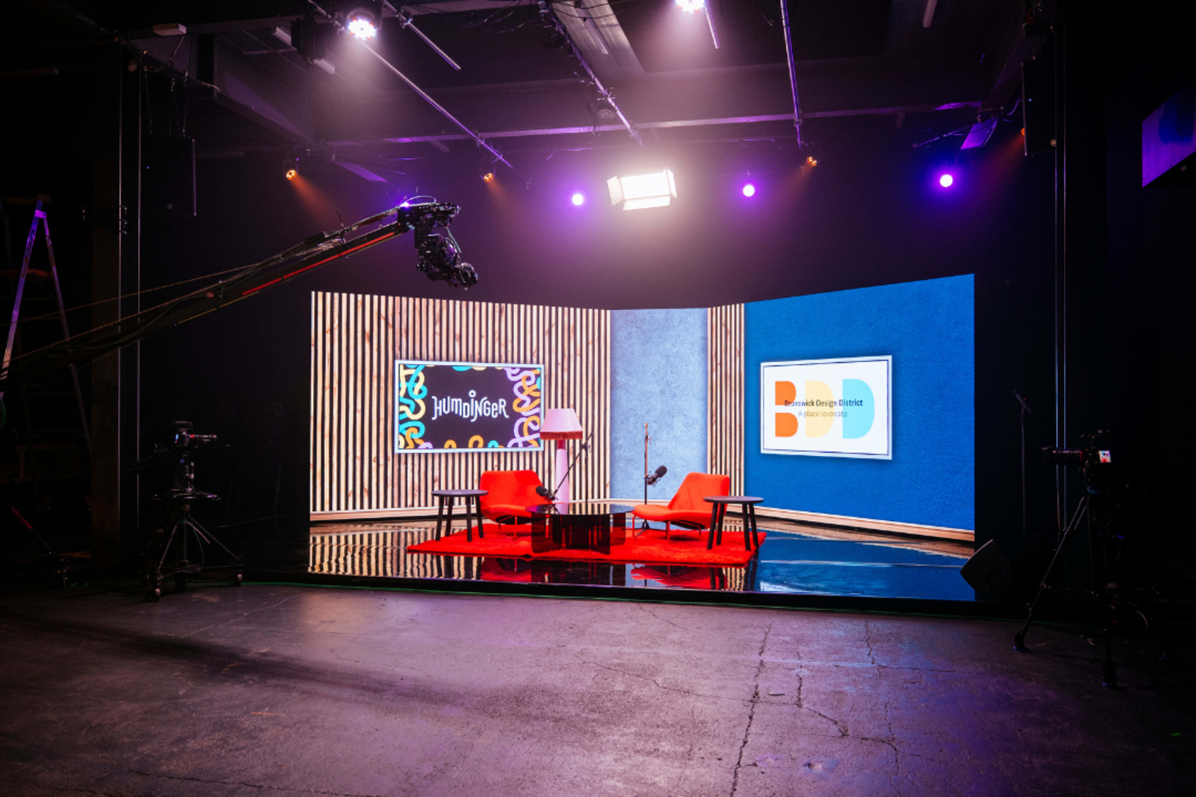 The image shows a modern studio setup featuring a pair of red chairs facing a round table, flanked by screens and colorful backdrop elements, with multiple cameras positioned around.