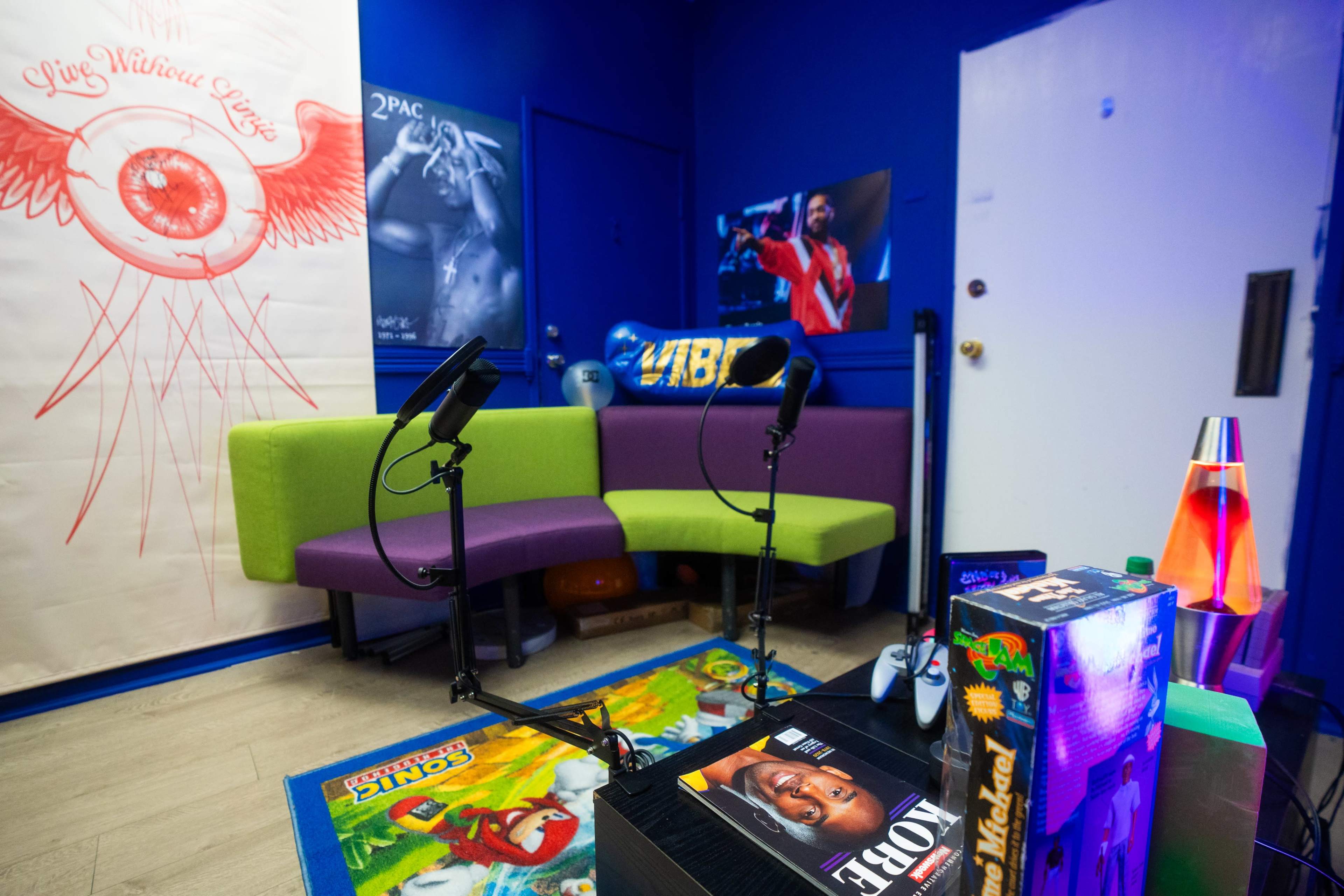 The image shows a colorful room with a green and purple sofa, two microphones on stands, a lava lamp, and various decorations, including a poster featuring 2Pac.