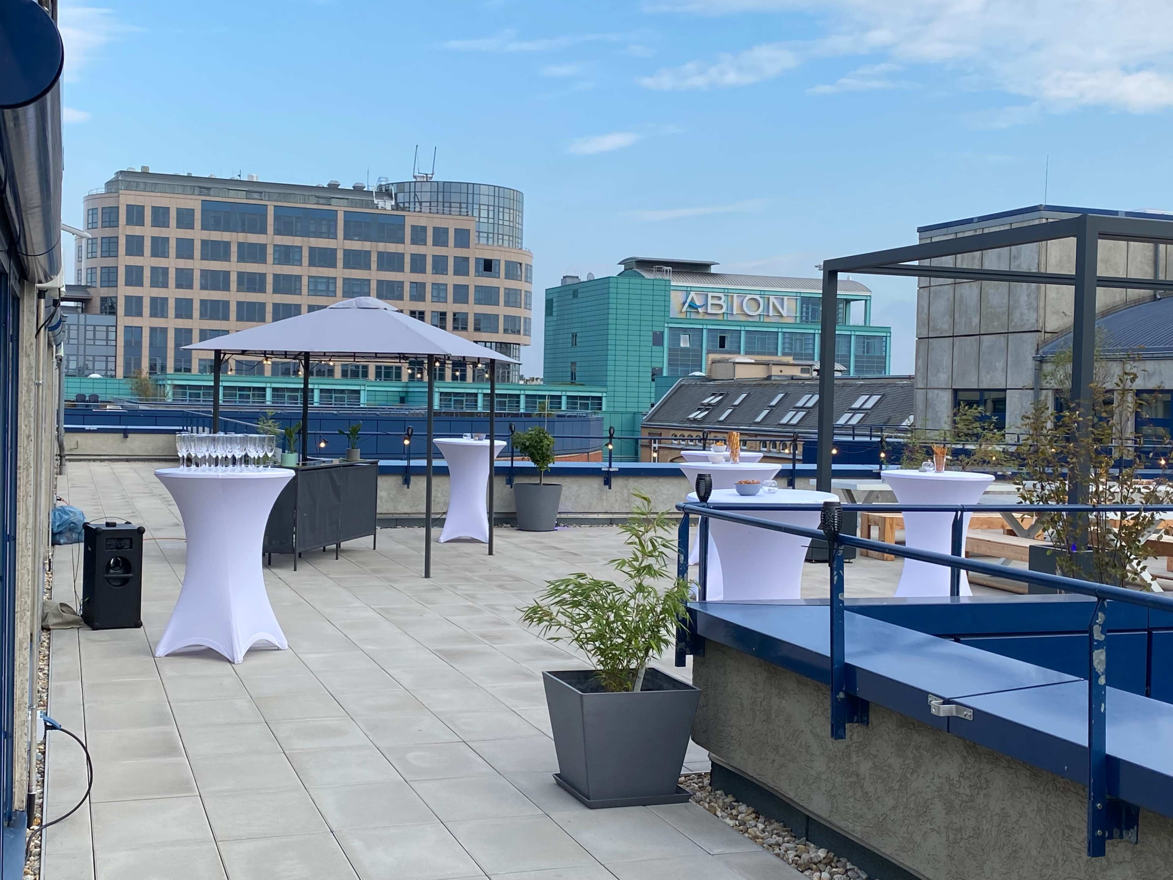The image shows a spacious rooftop event area featuring several circular tables with white tablecloths, a large canopy, and views of surrounding buildings.