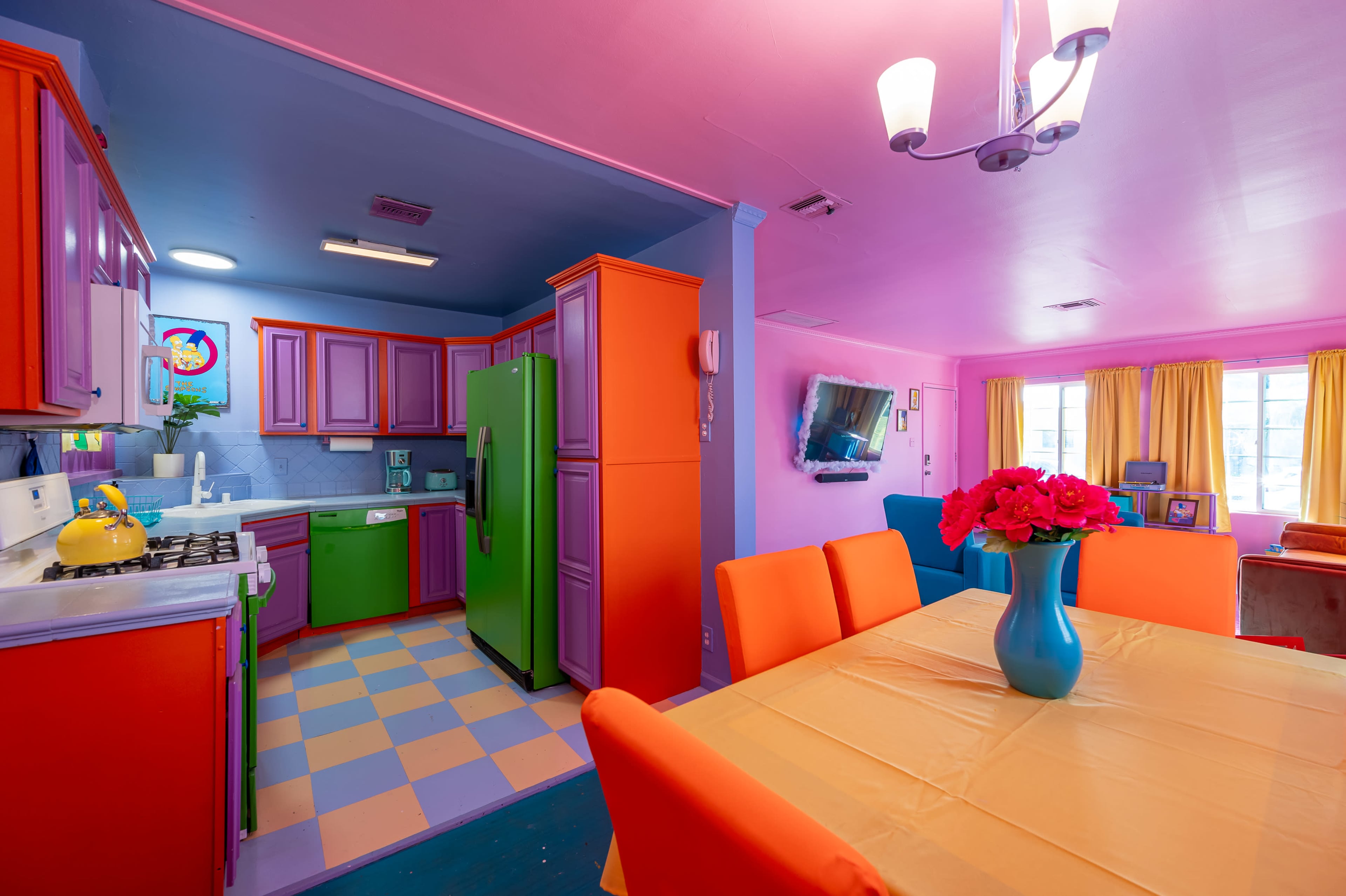 A brightly colored kitchen and dining area with orange and green cabinets, a pastel checkered floor, and a vibrant table set with flowers.