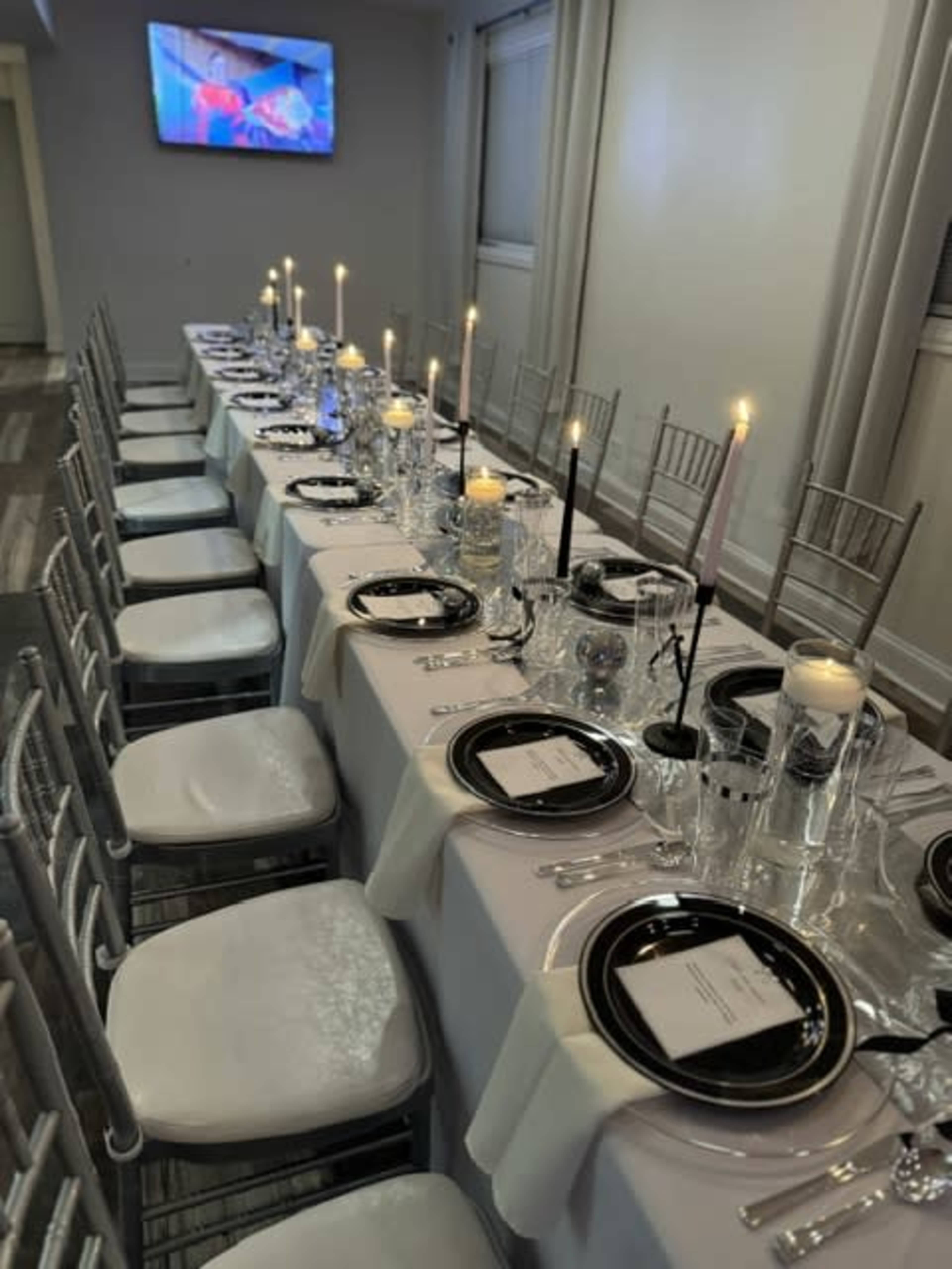 A long dining table is set with black plates, silver cutlery, and candles, arranged for a formal event, with a television screen in the background.