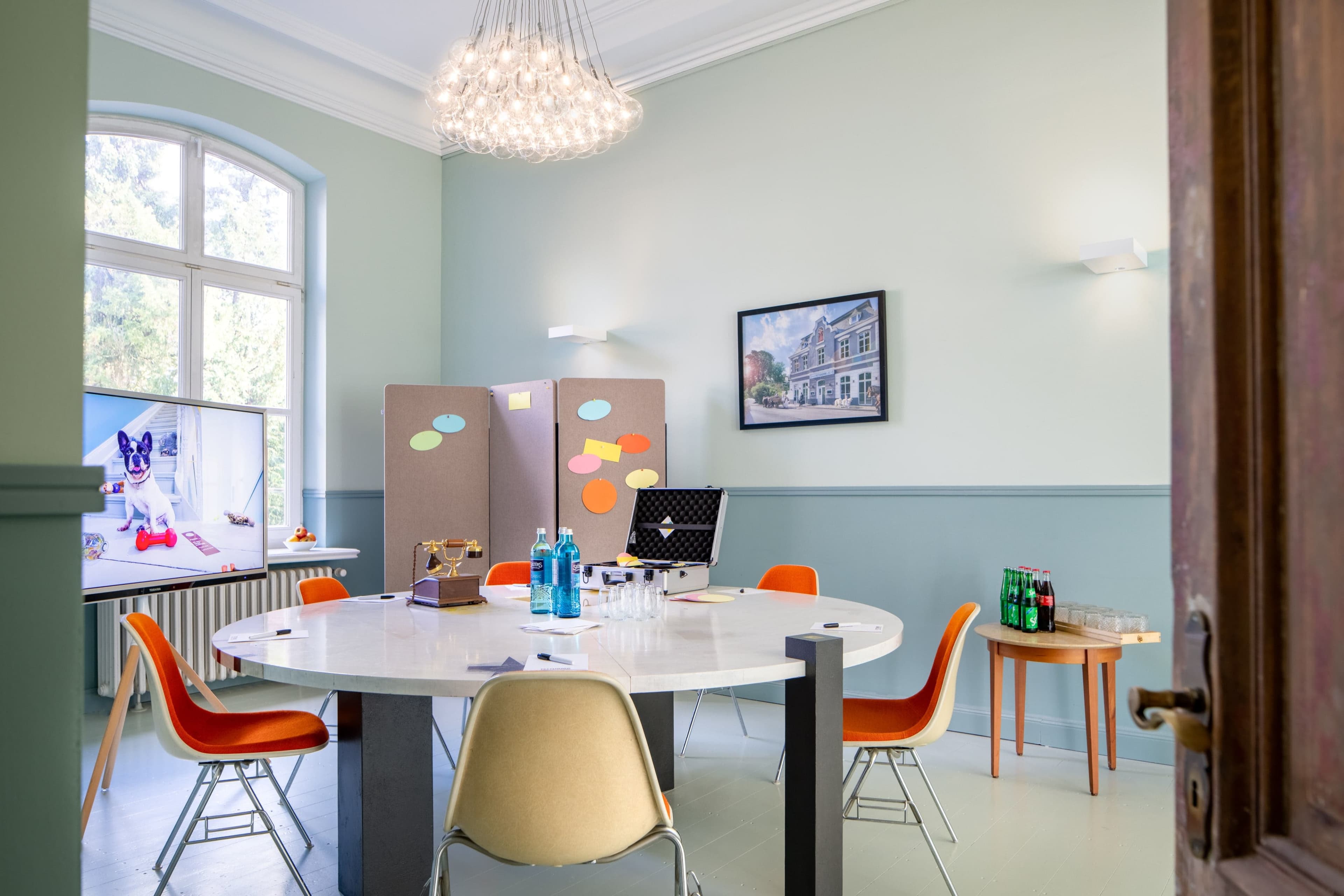 A conference room features a round table surrounded by orange chairs, with a large screen and a presentation board in the background.