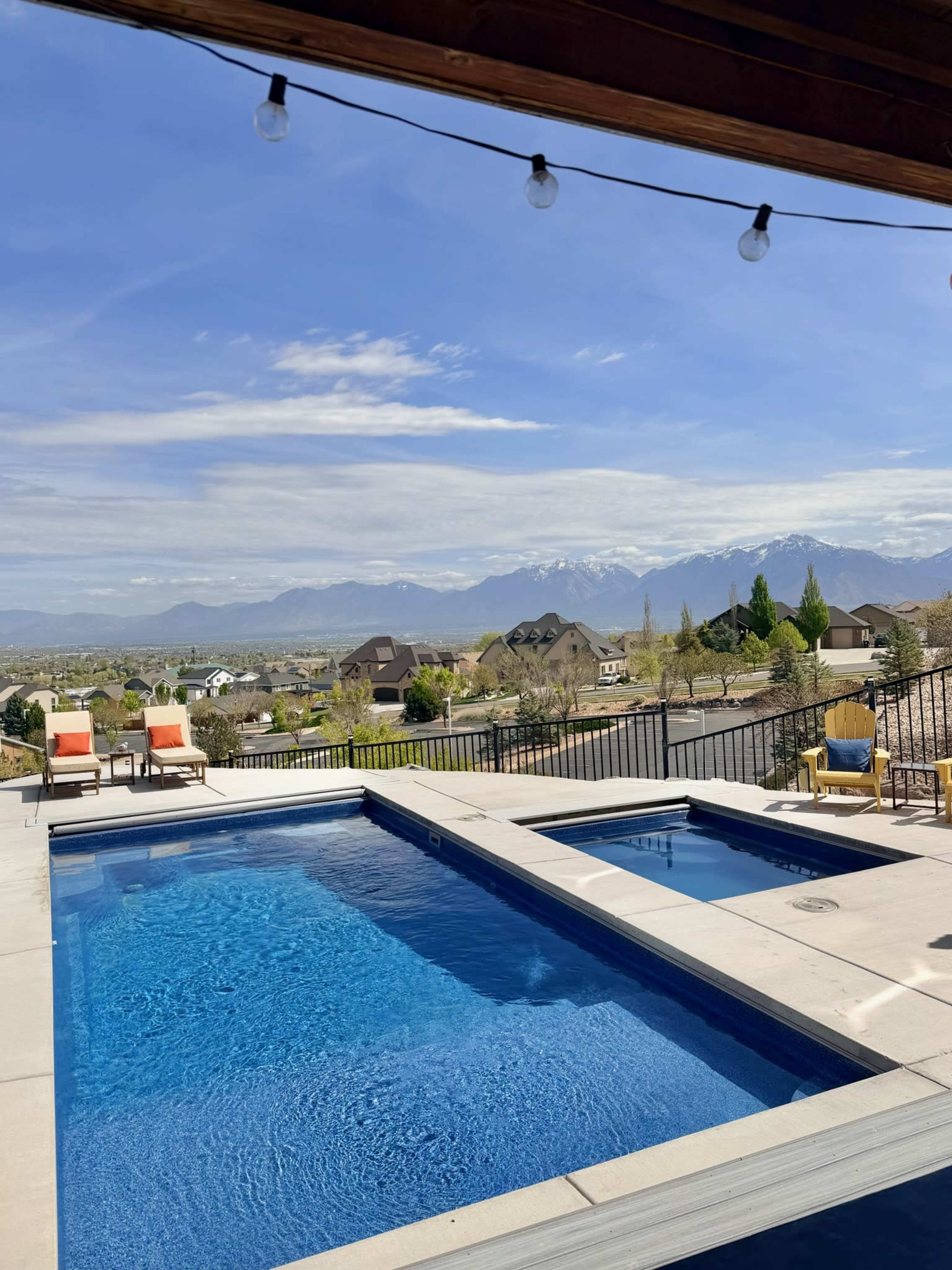 The image shows a rectangular swimming pool with a hot tub adjacent to it, set against a backdrop of mountains and houses under a clear blue sky.