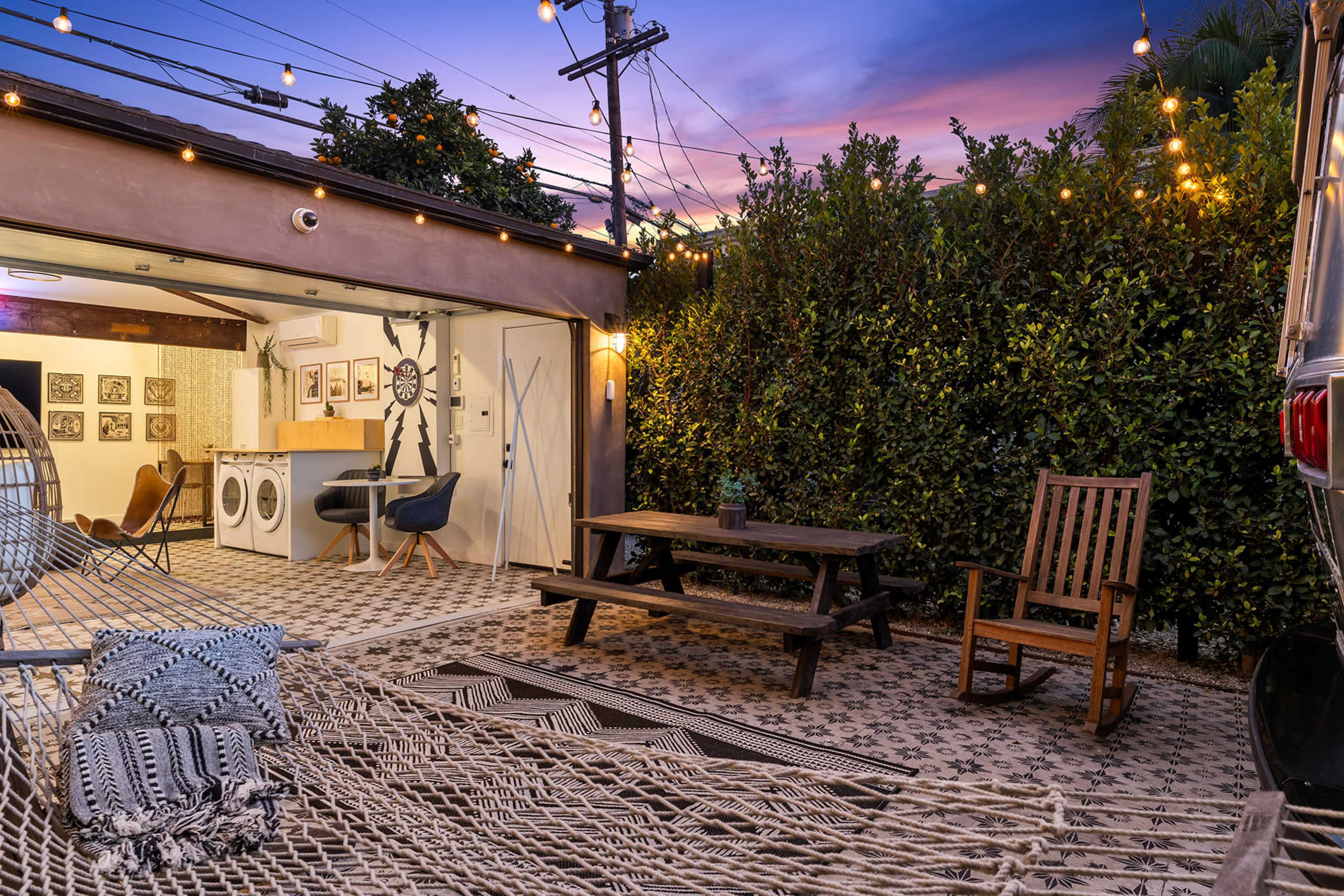 The image shows a cozy outdoor space featuring a hammock, a picnic table, and a laundry area with framed artwork in the background, illuminated by string lights at dusk.