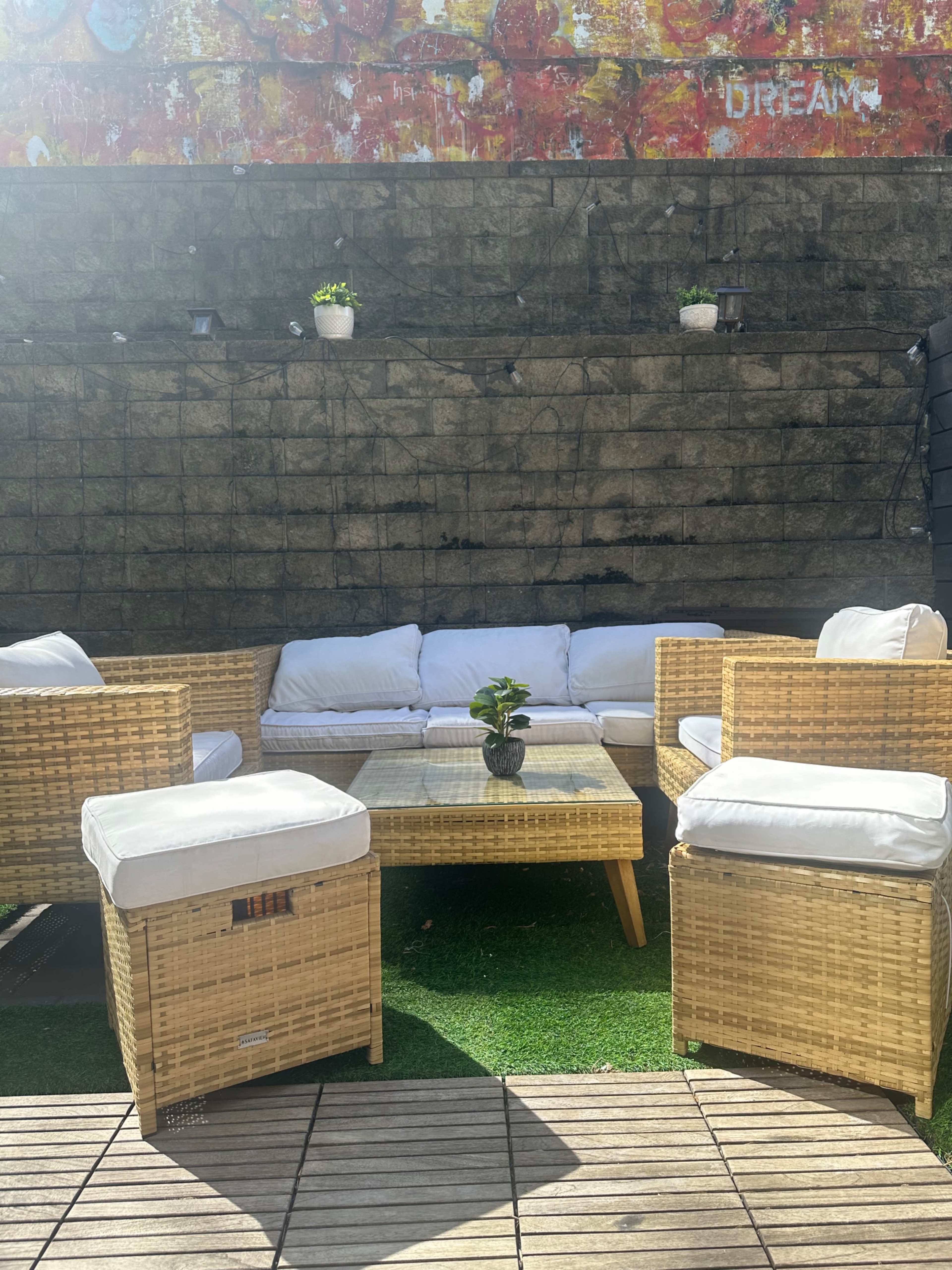 The image shows a cozy outdoor seating area with wicker furniture arranged around a square table on a green carpet.