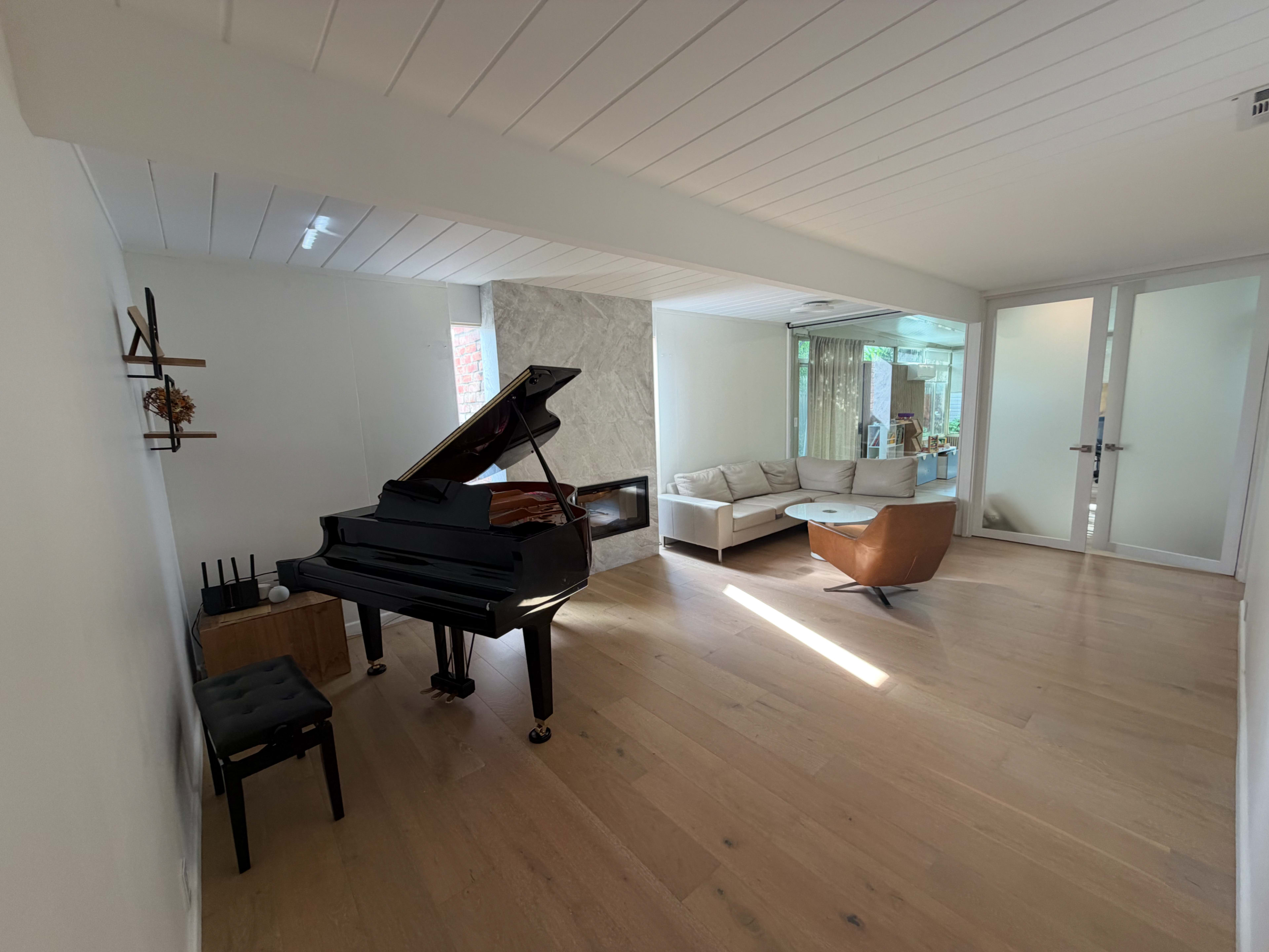 A grand piano sits beside a light-colored sofa set in a bright, spacious living area with large windows.