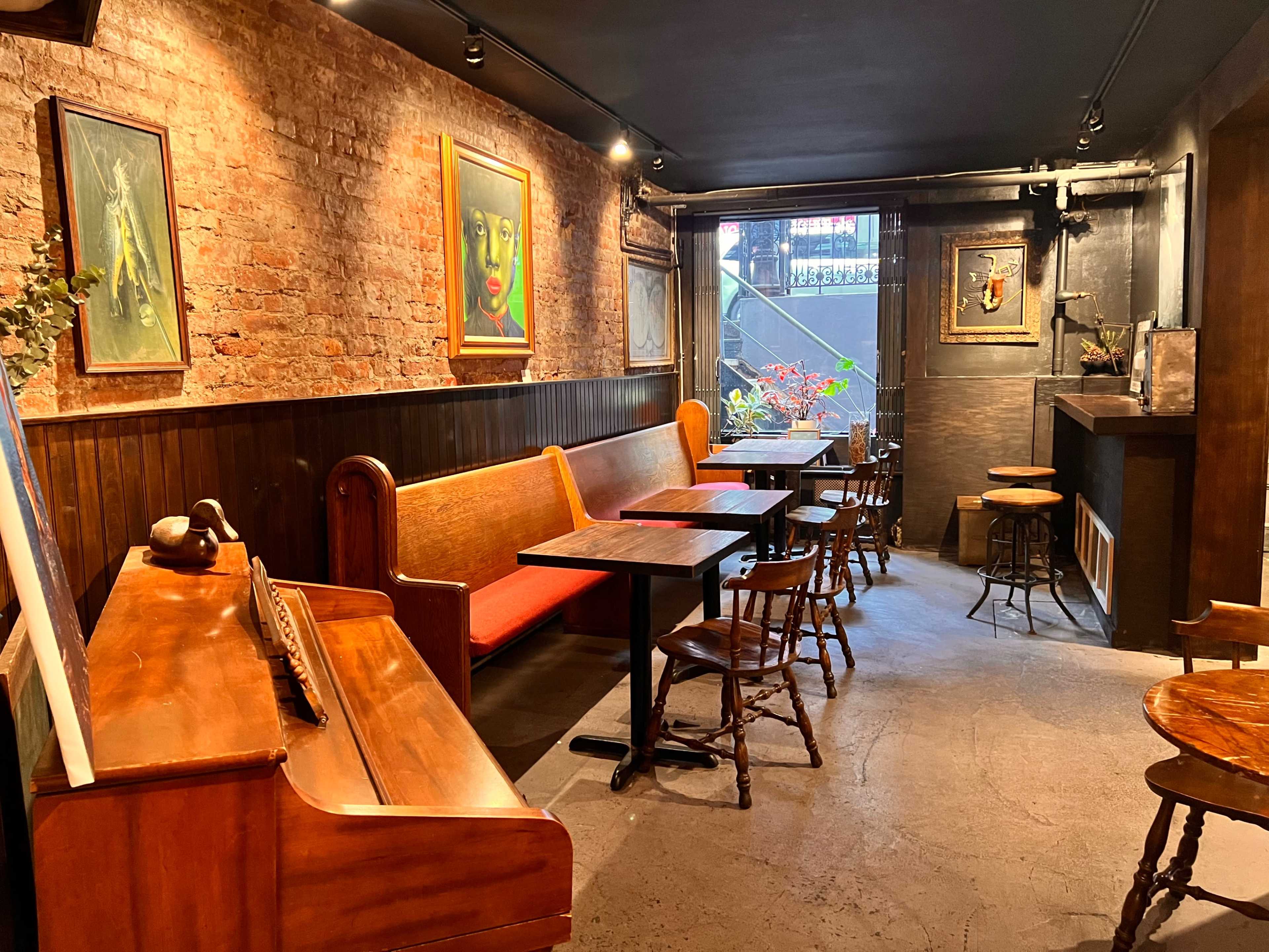 The image shows the interior of a cozy café featuring wooden tables, chairs, and a vintage piano against a brick wall with framed artwork.