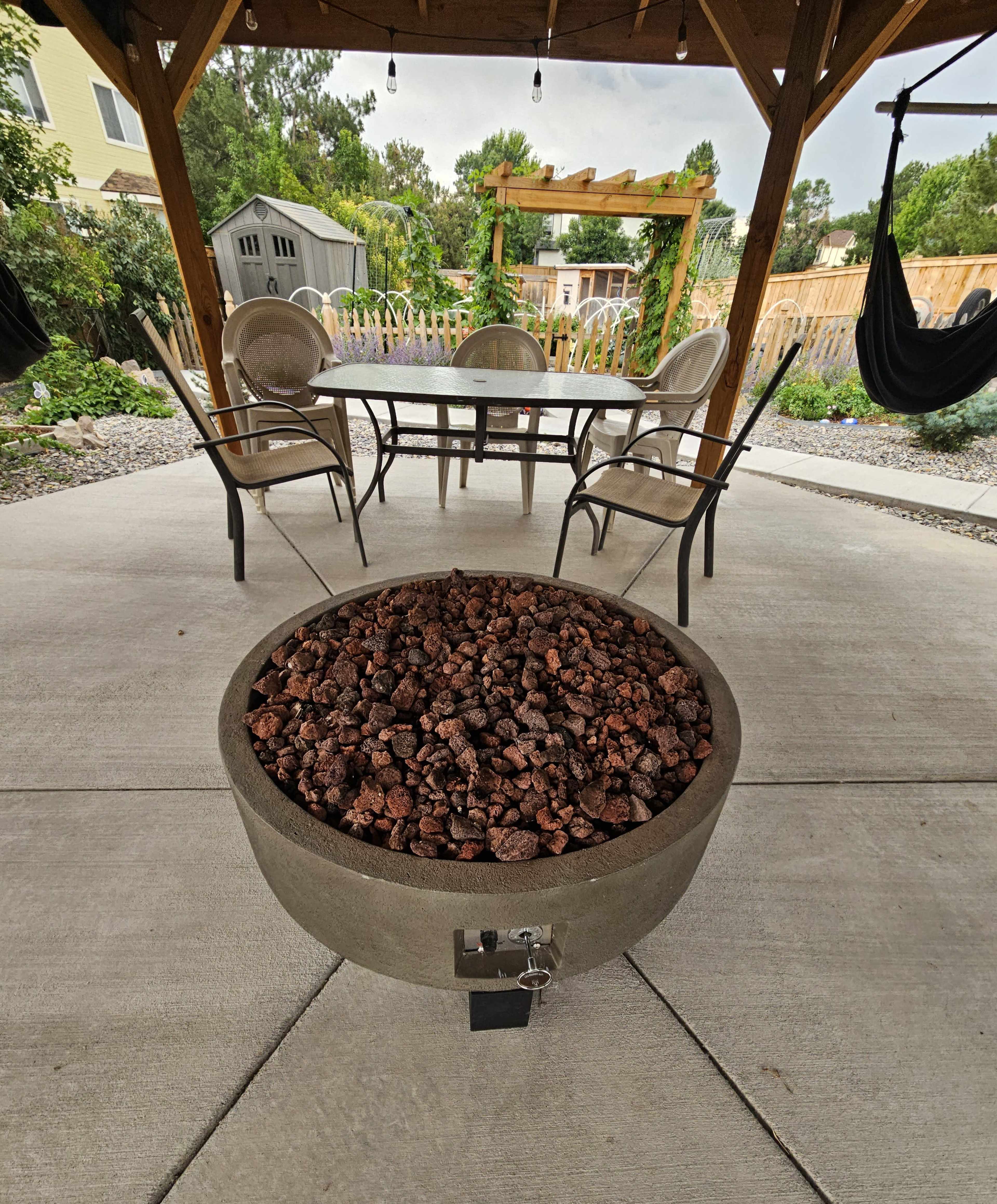 A round fire pit filled with rocks is centered under a covered patio with chairs and a table in the background.