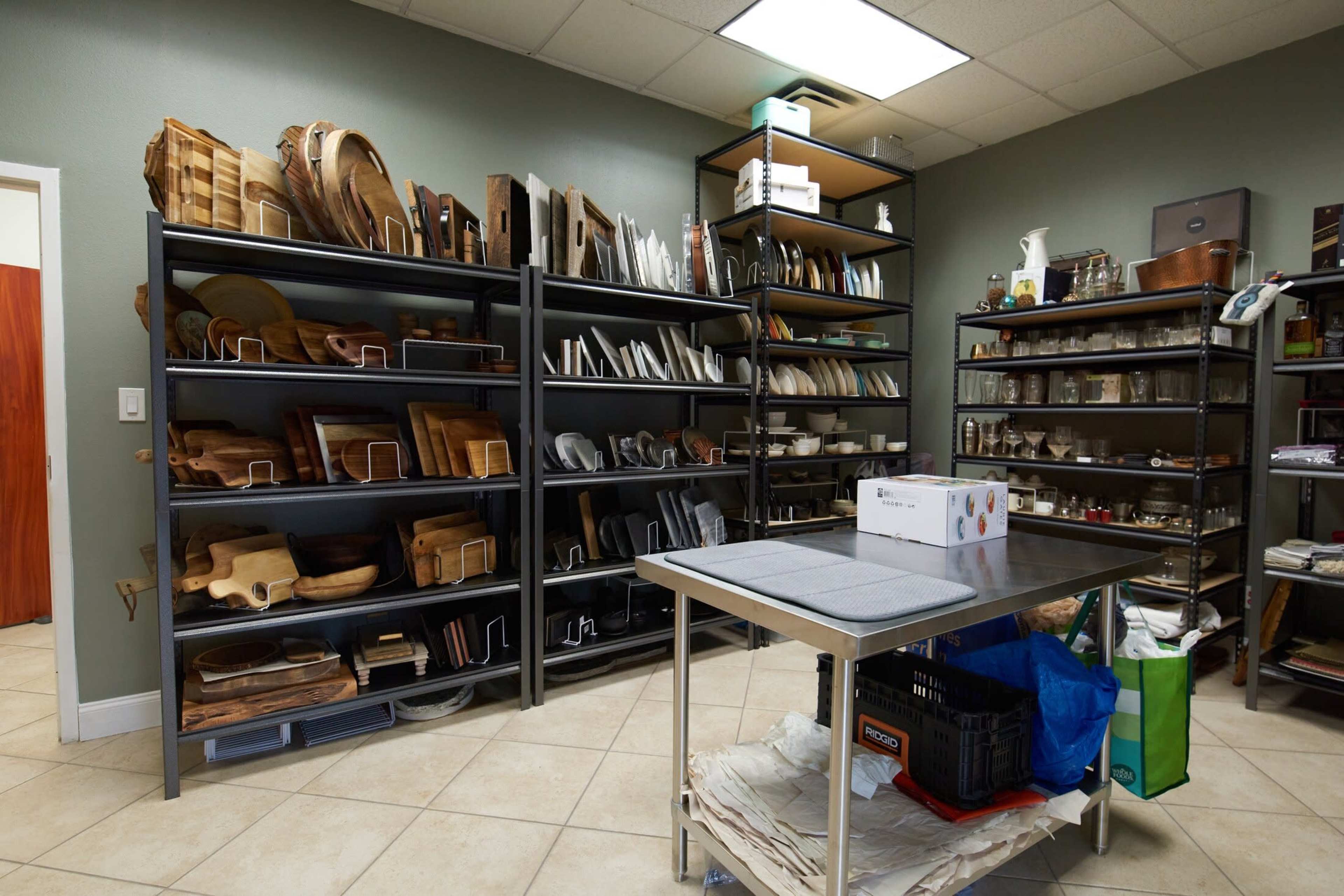 A well-organized storage room features metal shelves filled with wooden and ceramic kitchenware, alongside a table in the center.