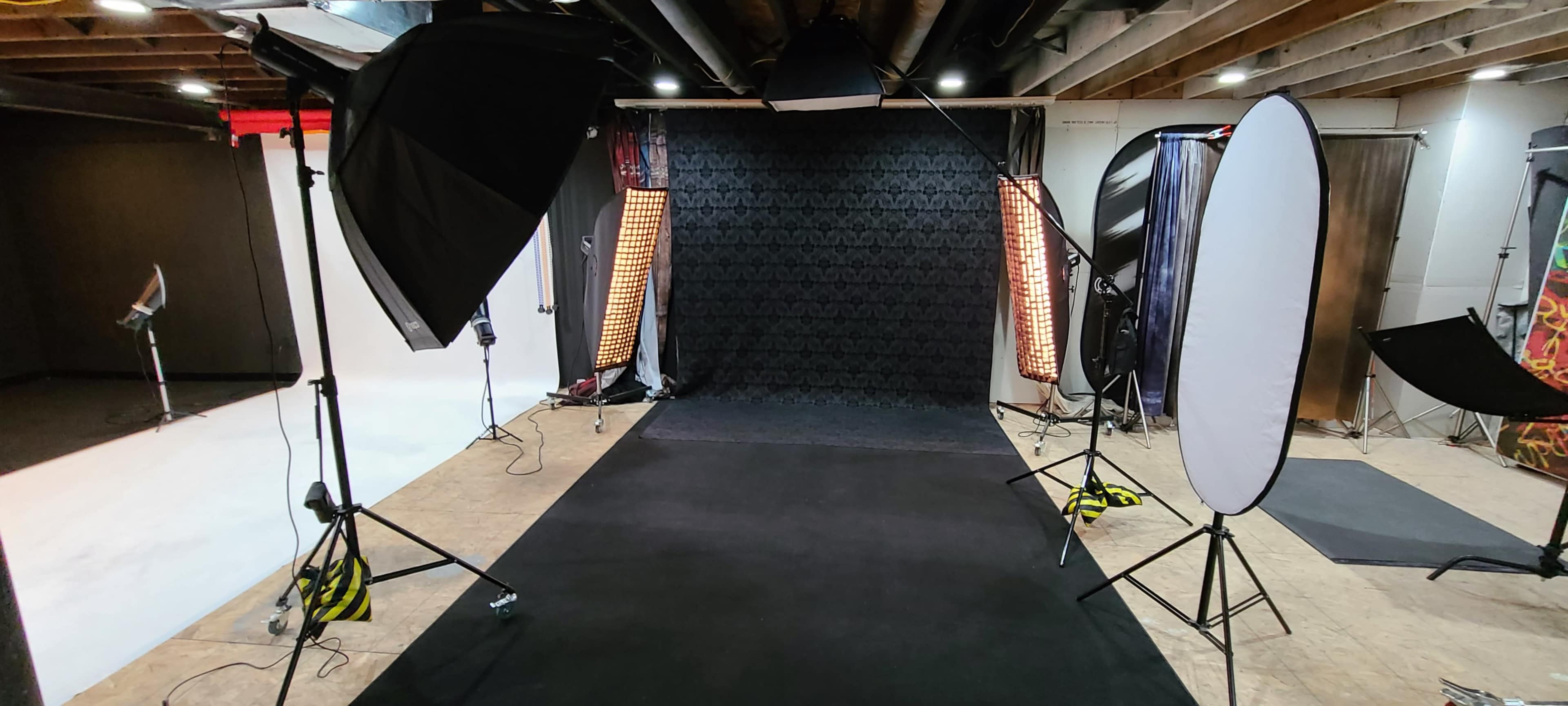 A photography studio setup features a black backdrop, multiple lighting equipment, and reflectors arranged around a black carpeted area.