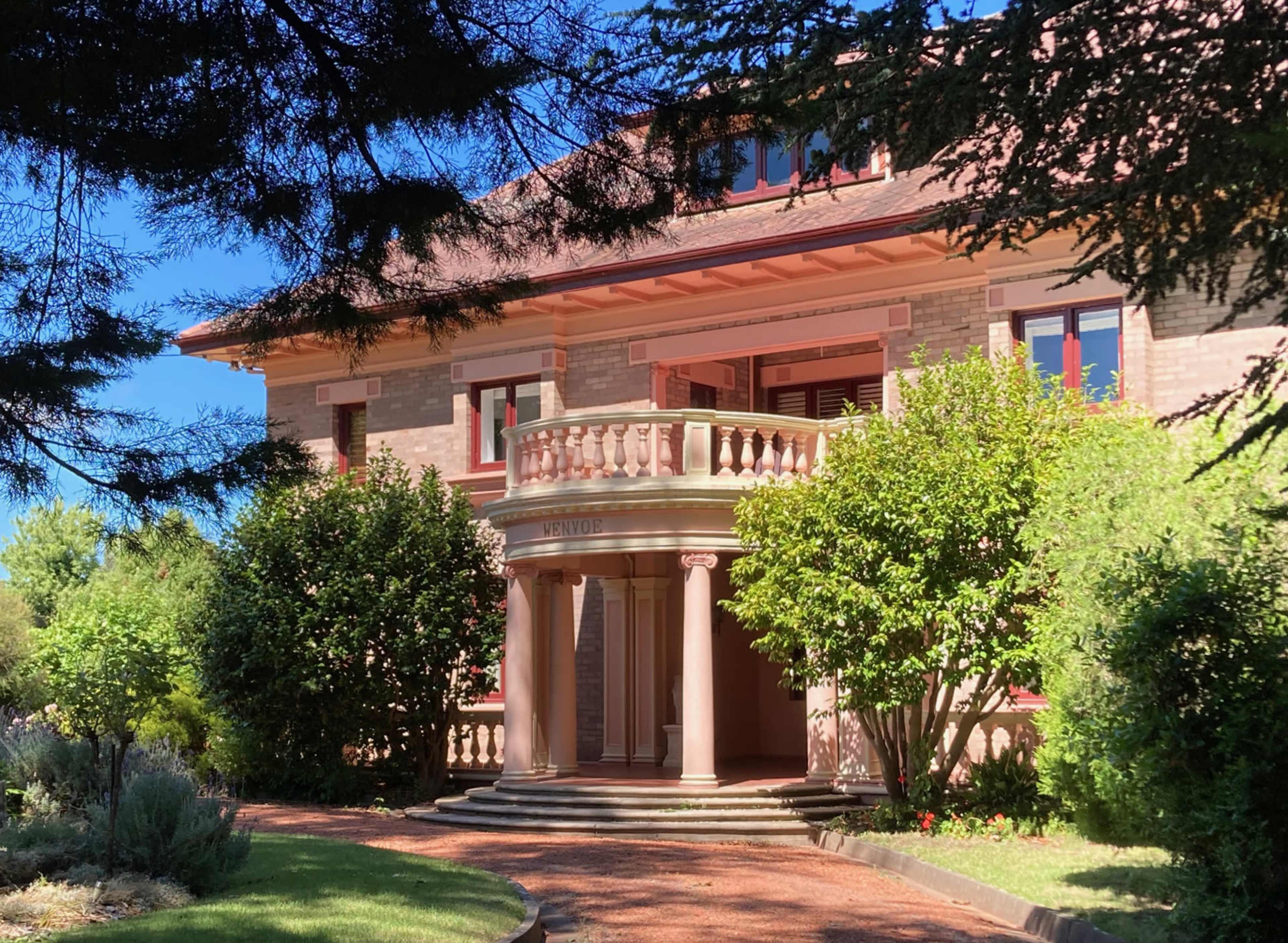 A grand, two-story brick house with a front porch featuring columns and surrounded by well-kept greenery.