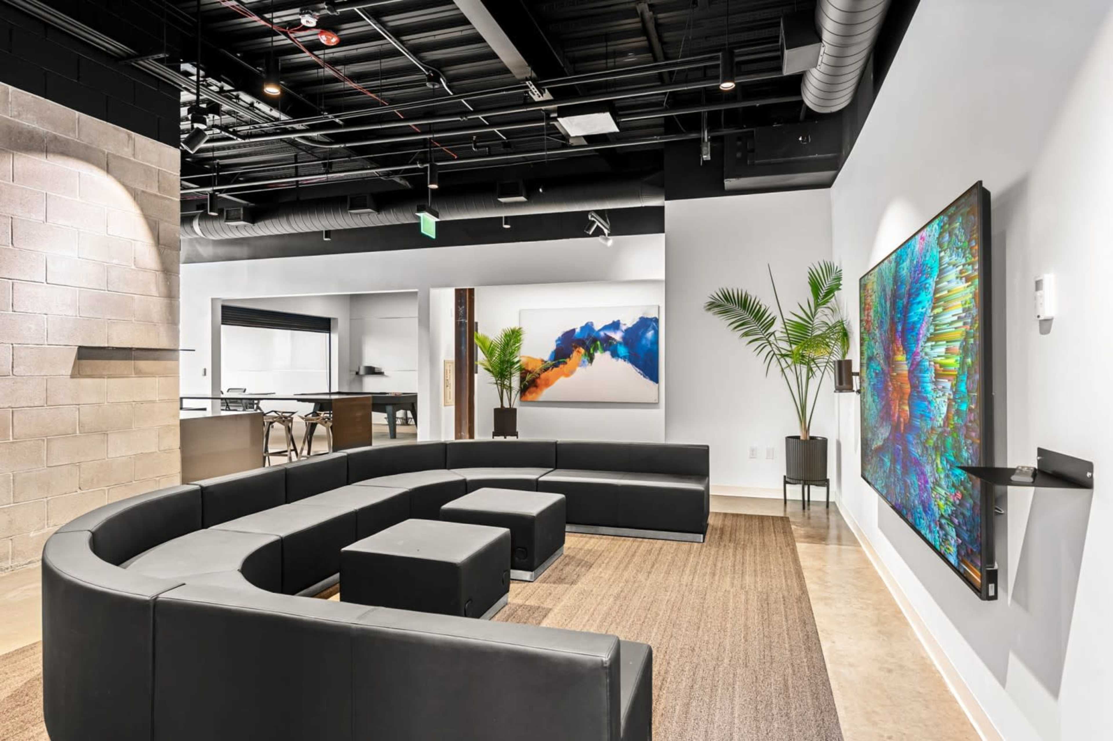 The image shows a modern lounge area featuring a curved black seating arrangement, a large colorful artwork on the wall, and a kitchen space visible in the background.