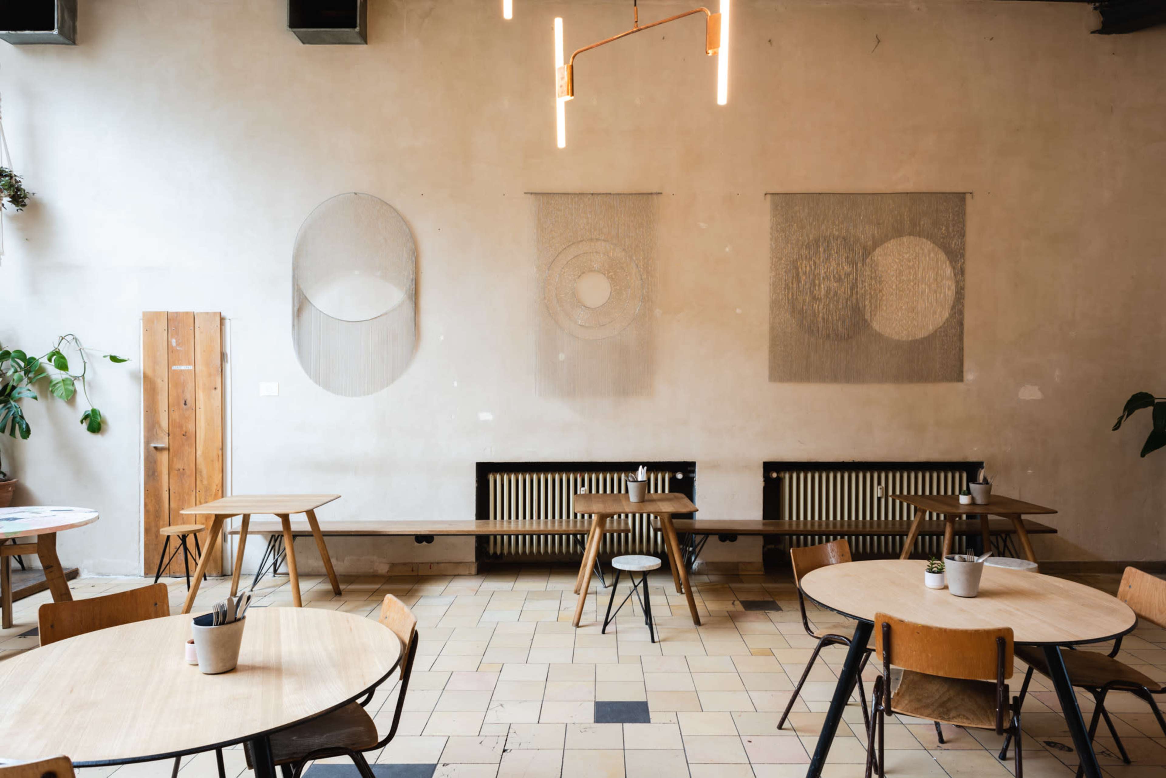 A minimalist café interior with wooden tables and chairs, a long bench along the wall, and geometric wall hangings above a radiator.