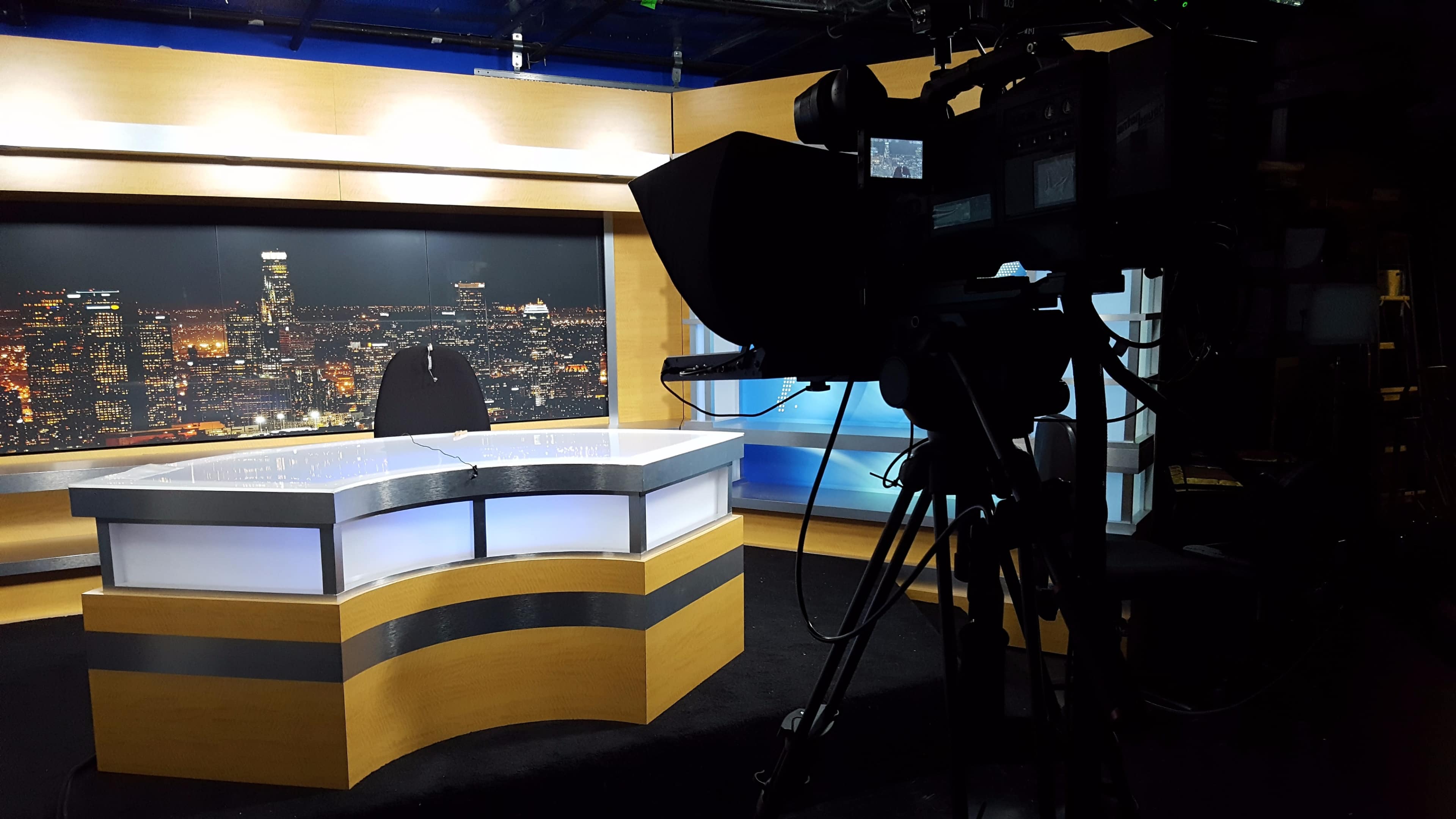 A television studio with a news desk and a camera positioned to capture a city skyline backdrop at night.