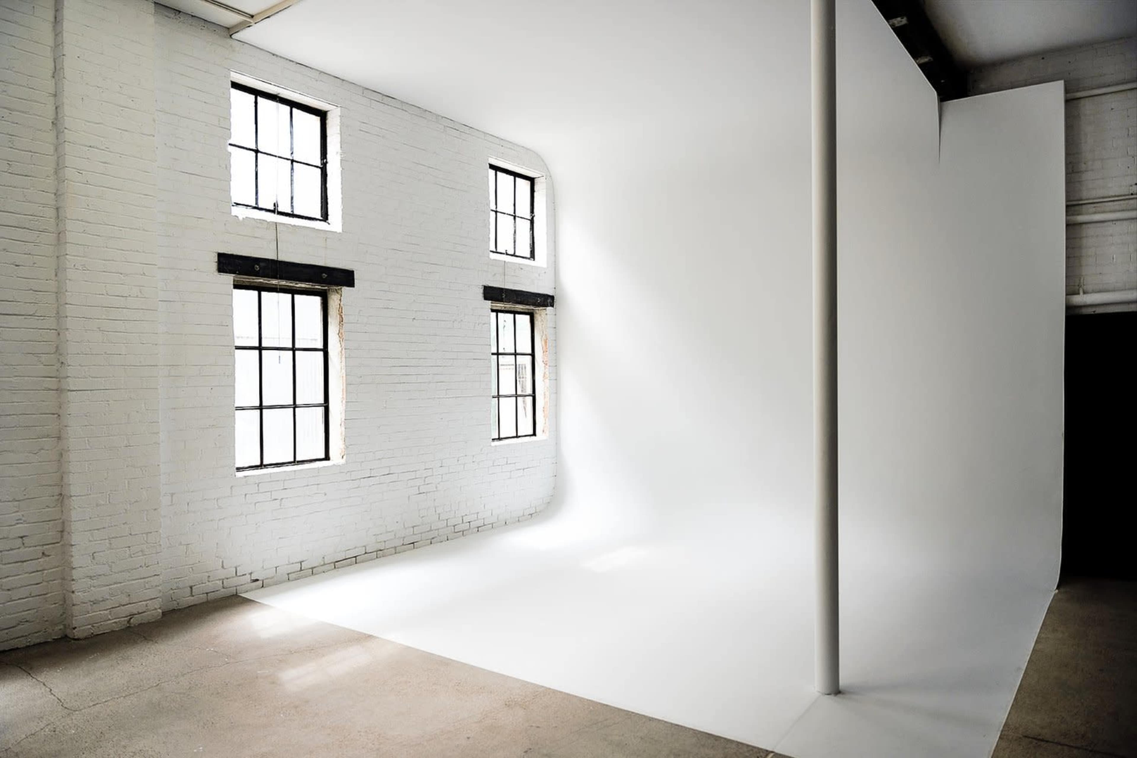A well-lit photography studio featuring white brick walls, large windows, and a seamless white backdrop.
