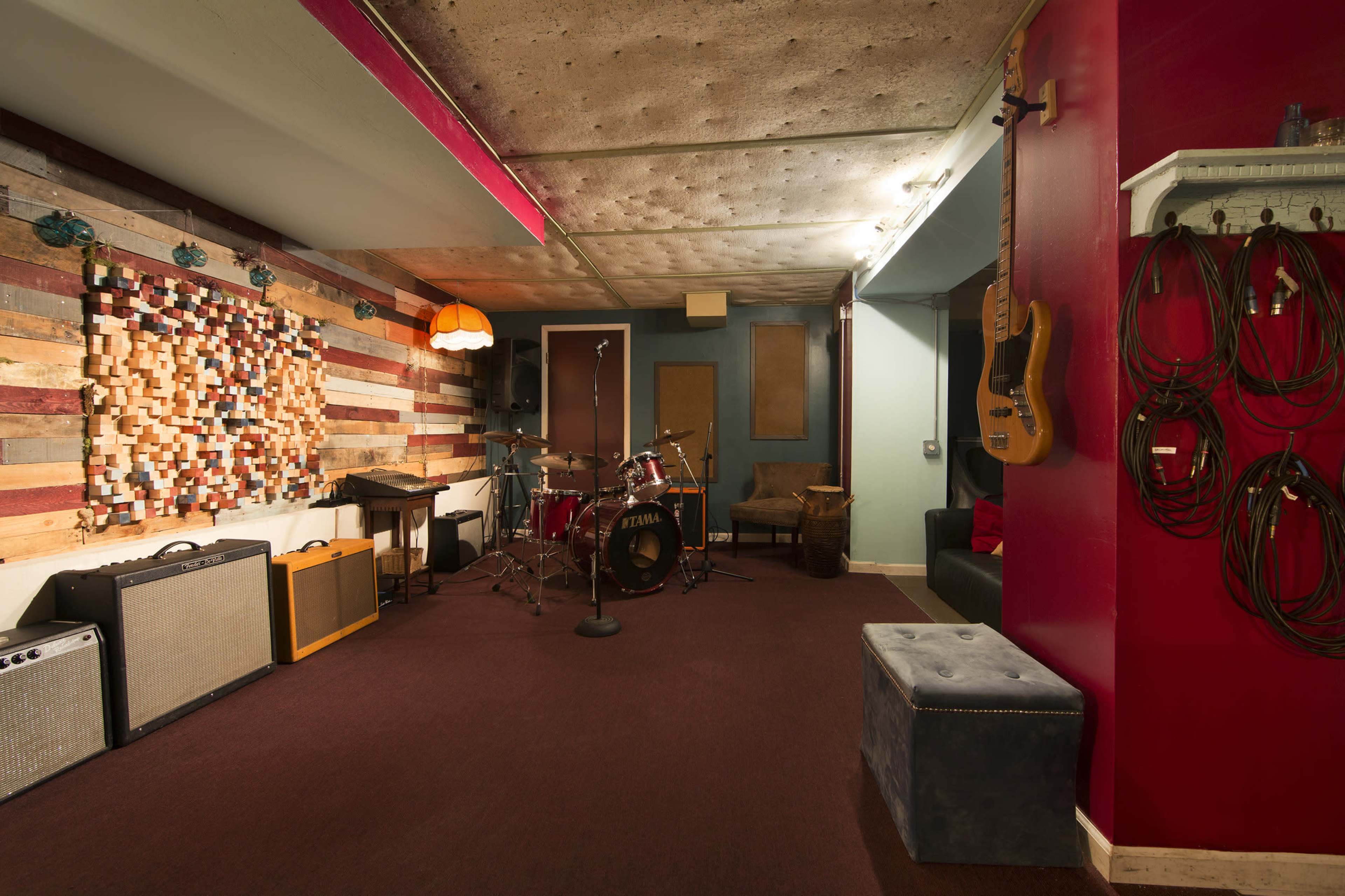 A music practice space featuring a drum set, amplifiers, a wooden wall made of colorful planks, and a cozy seating area.